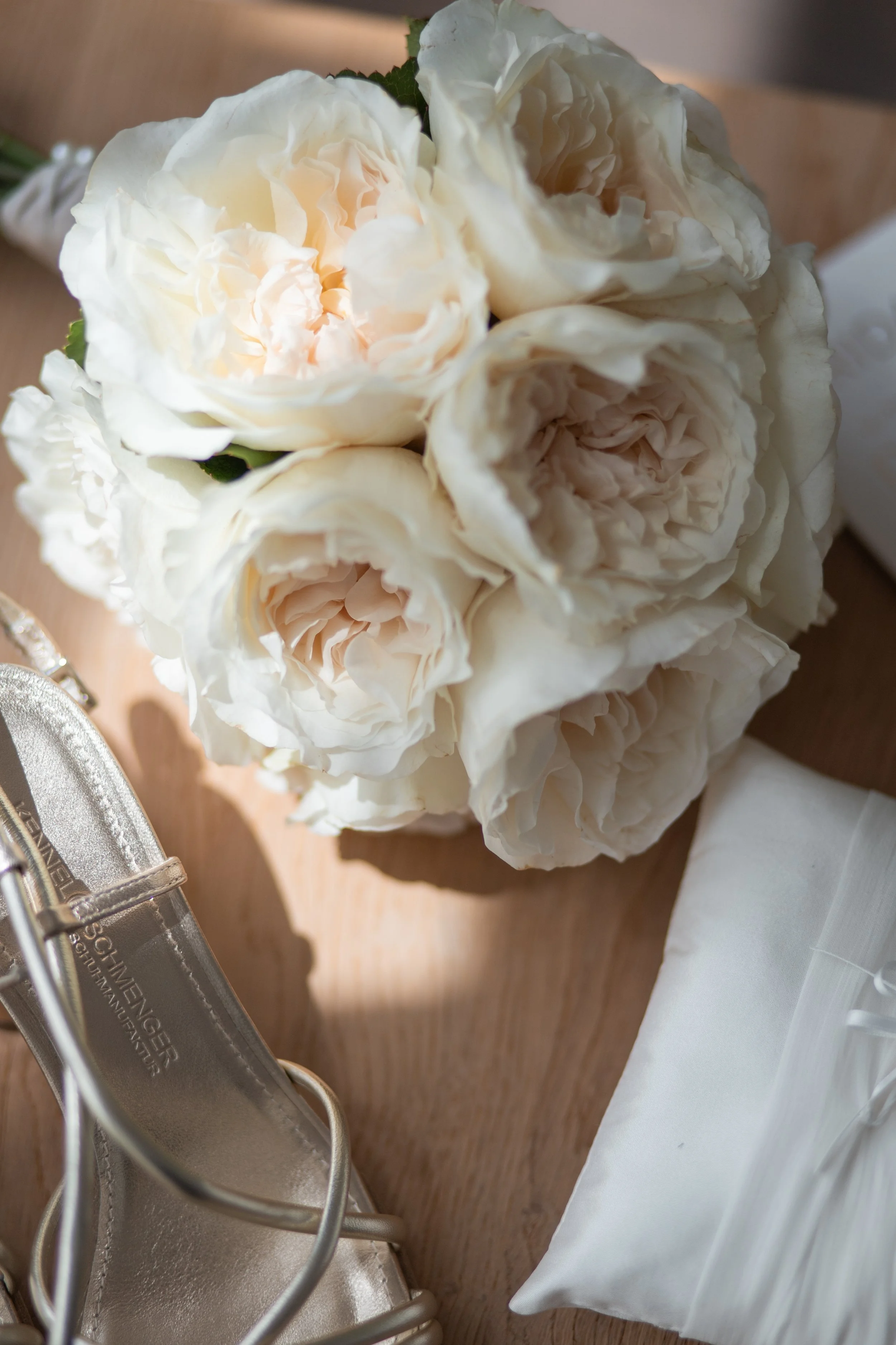 A pair of silver high-heeled shoes next to a white fabric, a bouquet of cream-colored roses, a white cloth, and a wooden surface.