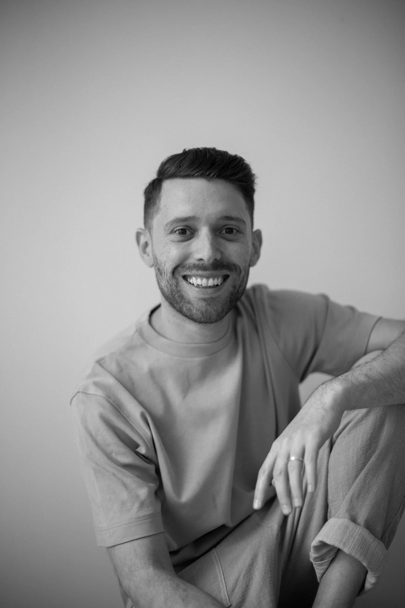 Black and white photo of a smiling man with short hair and beard, wearing a long sleeve shirt, sitting with arms crossed in front of him against a plain background.