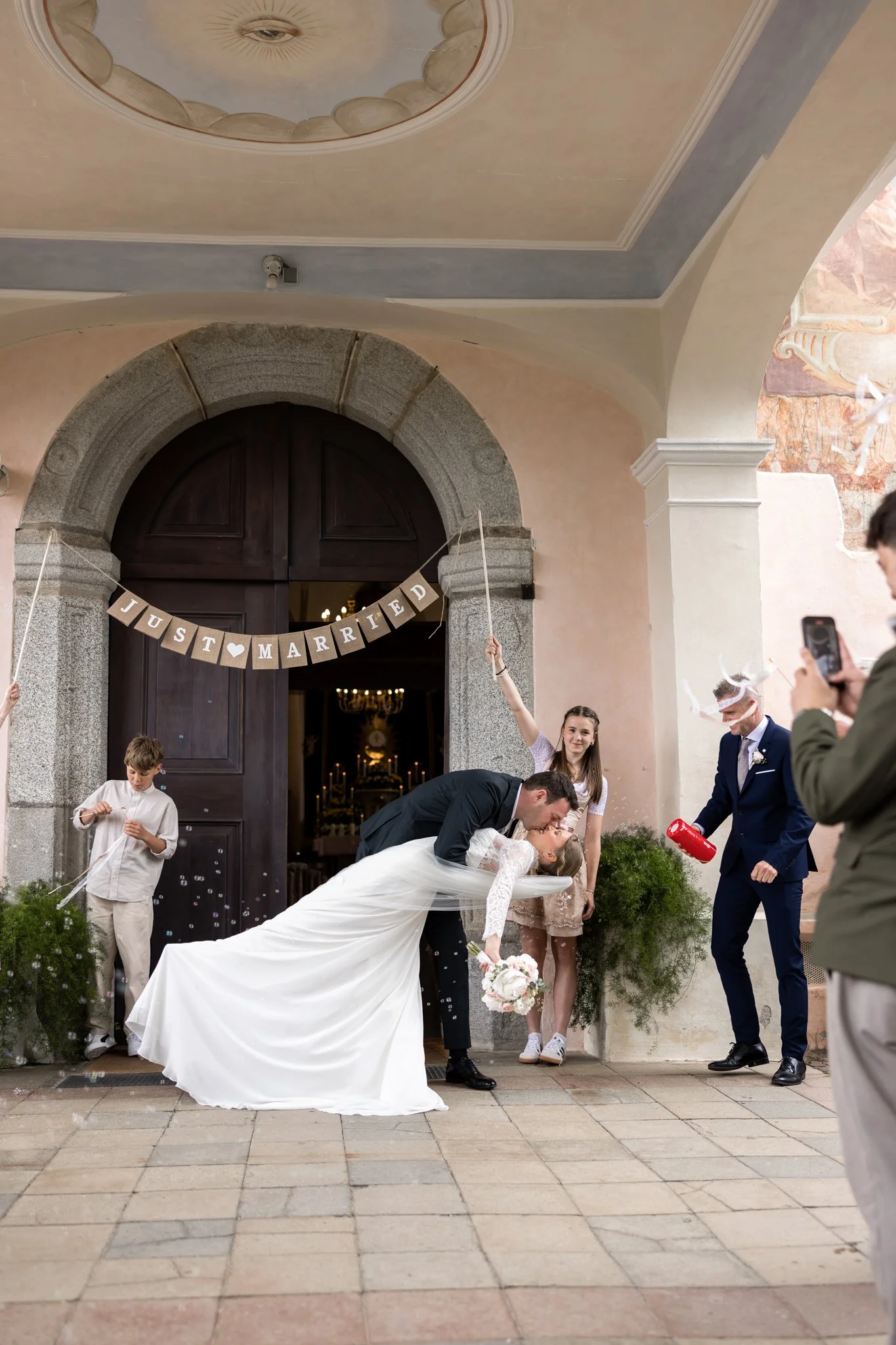 A bride and groom sharing a kiss outside a church after their wedding, with friends and family celebrating around them, and a 'Just Married' banner hanging over the door.