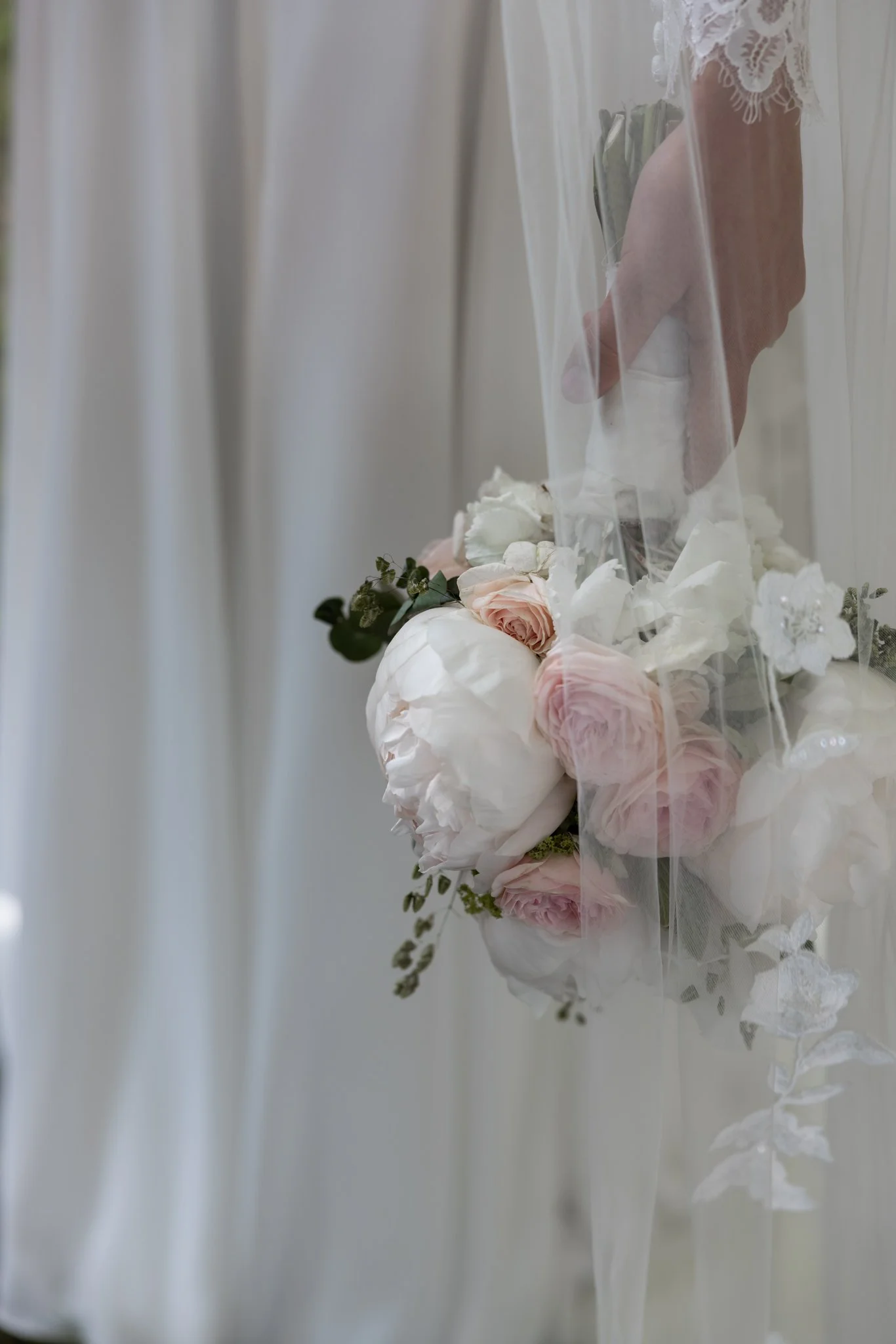 Close-up of a person's hand holding a bouquet of white and blush pink flowers, seen through a wedding veil.
