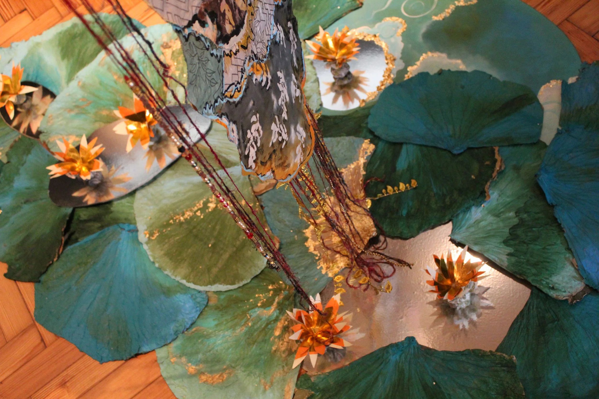 Decorative water lilies with large green leaves, paper lotus flowers, and hanging beads on a reflective surface, with a wooden floor underneath.
