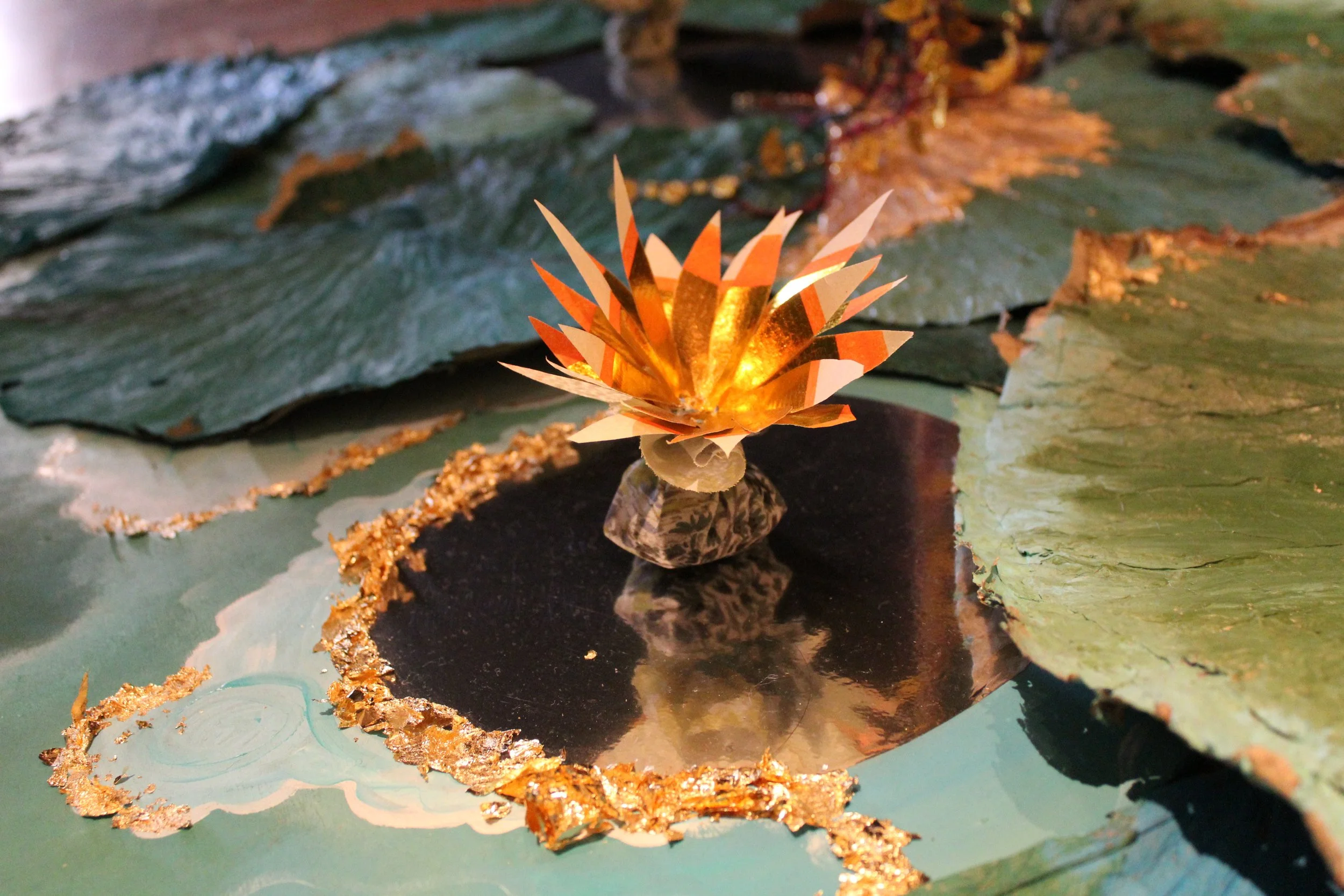 A decorative paper lotus flower with gold accents floating on a reflective surface, surrounded by green leaves with gold-trimmed edges.