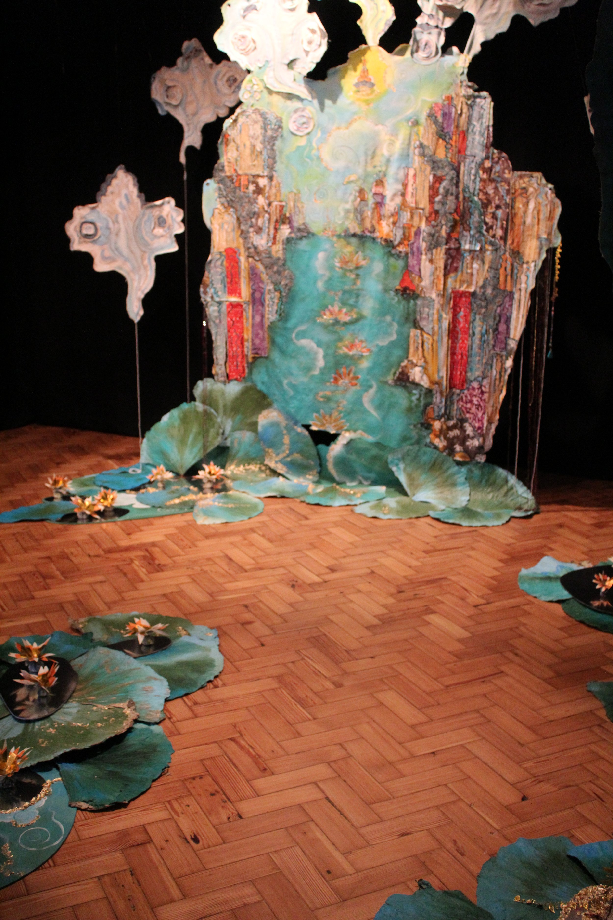 An art installation depicting a stylized landscape with a river, floating flowers, and large lily pads, with paper clouds hanging overhead, on a parquet floor.