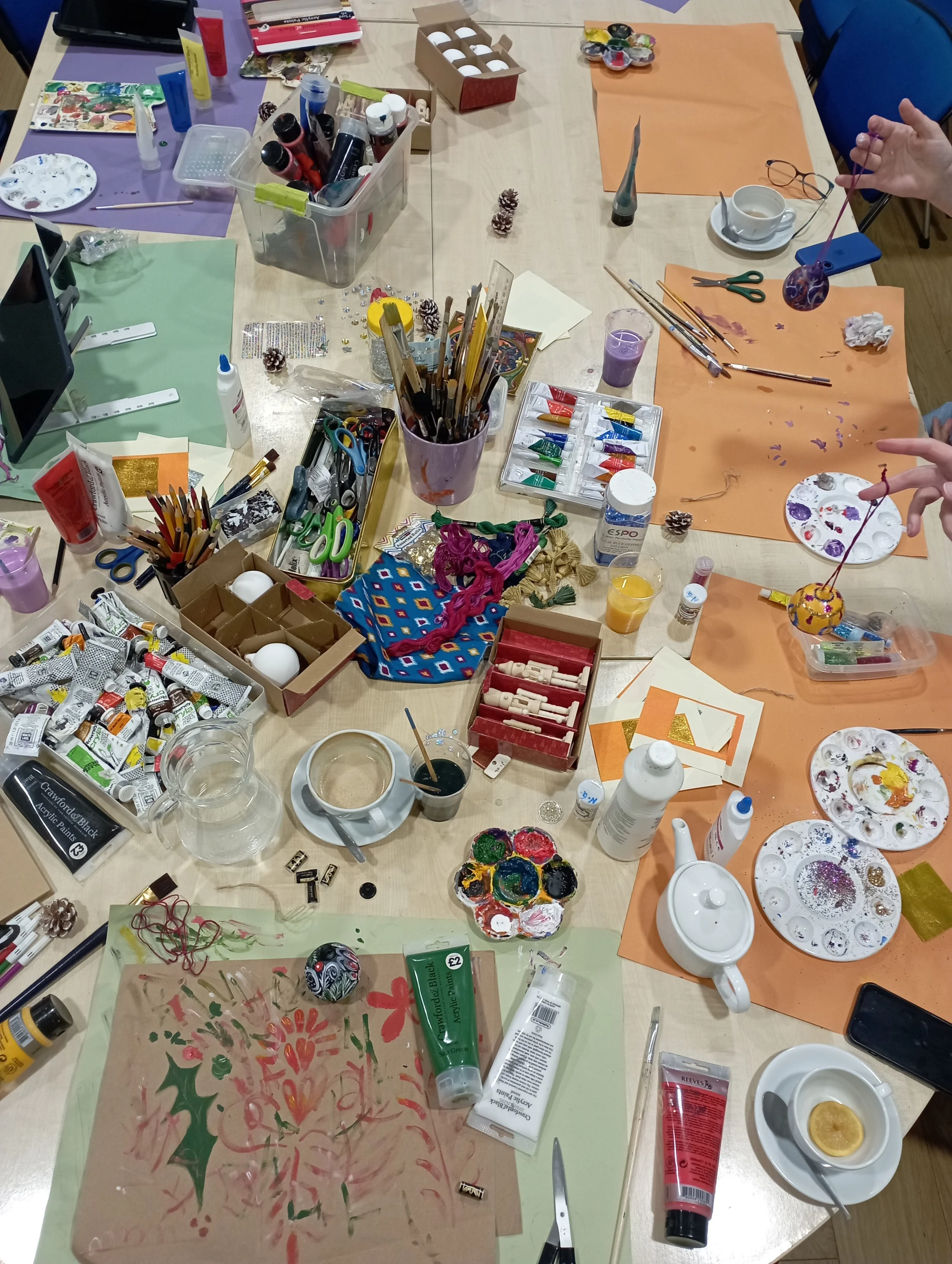 A cluttered table with art supplies including paints, brushes, scissors, glue, paper, and decorative items, used for crafting or painting.