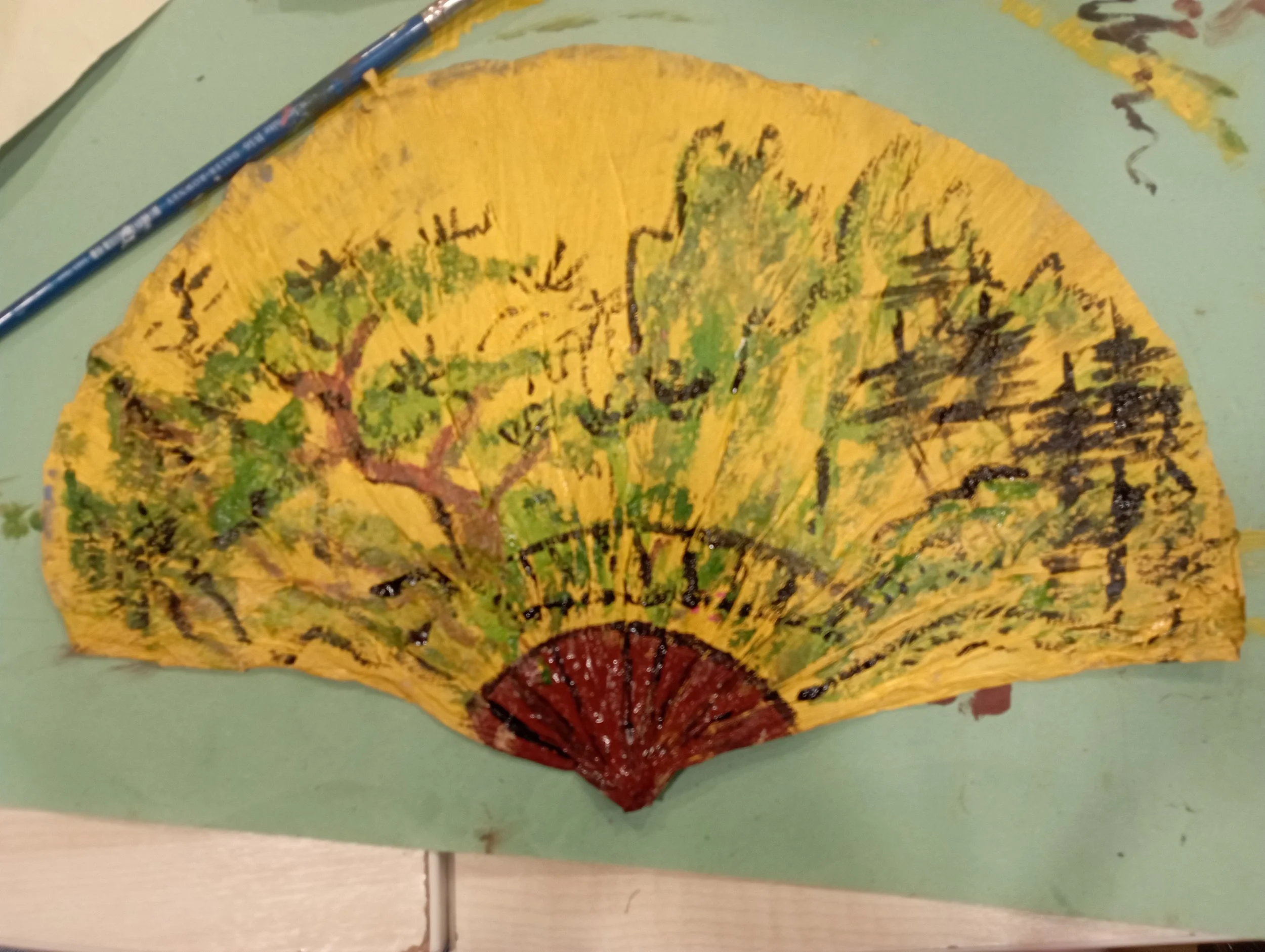 Colorful hand-painted paper fan featuring a landscape with trees on a yellow background.