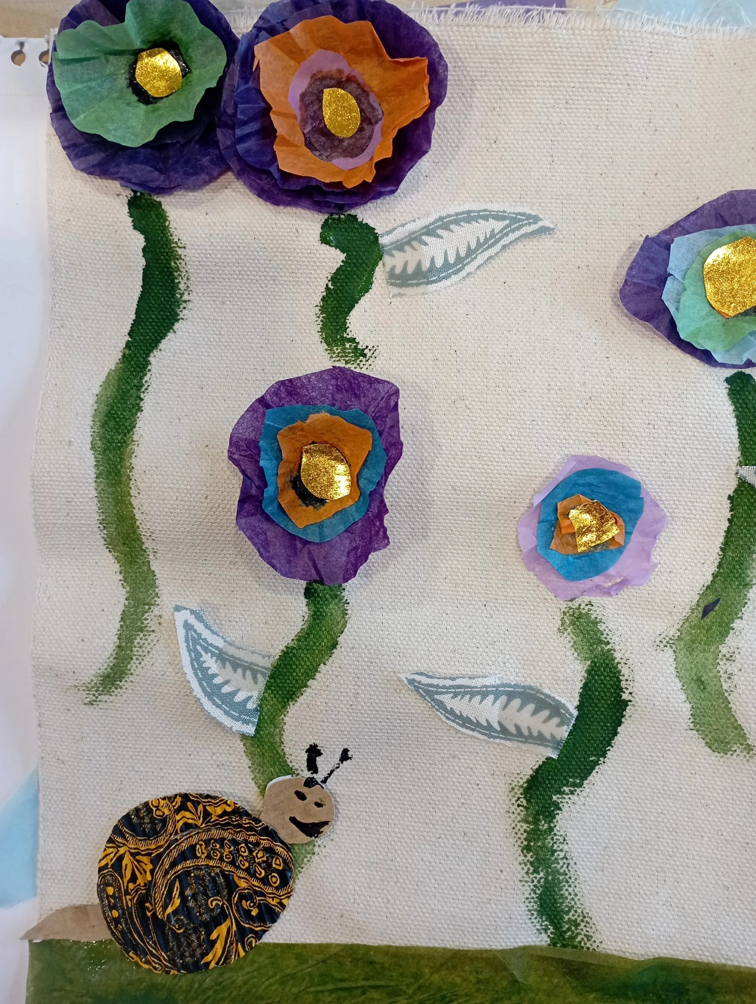 A craft artwork featuring paper flowers with purple, orange, blue, and green petals and gold centers, green textured stems, gray and white leaves, and a butterfly with intricate yellow, black, and blue patterns on its wings, all arranged on a fabric 