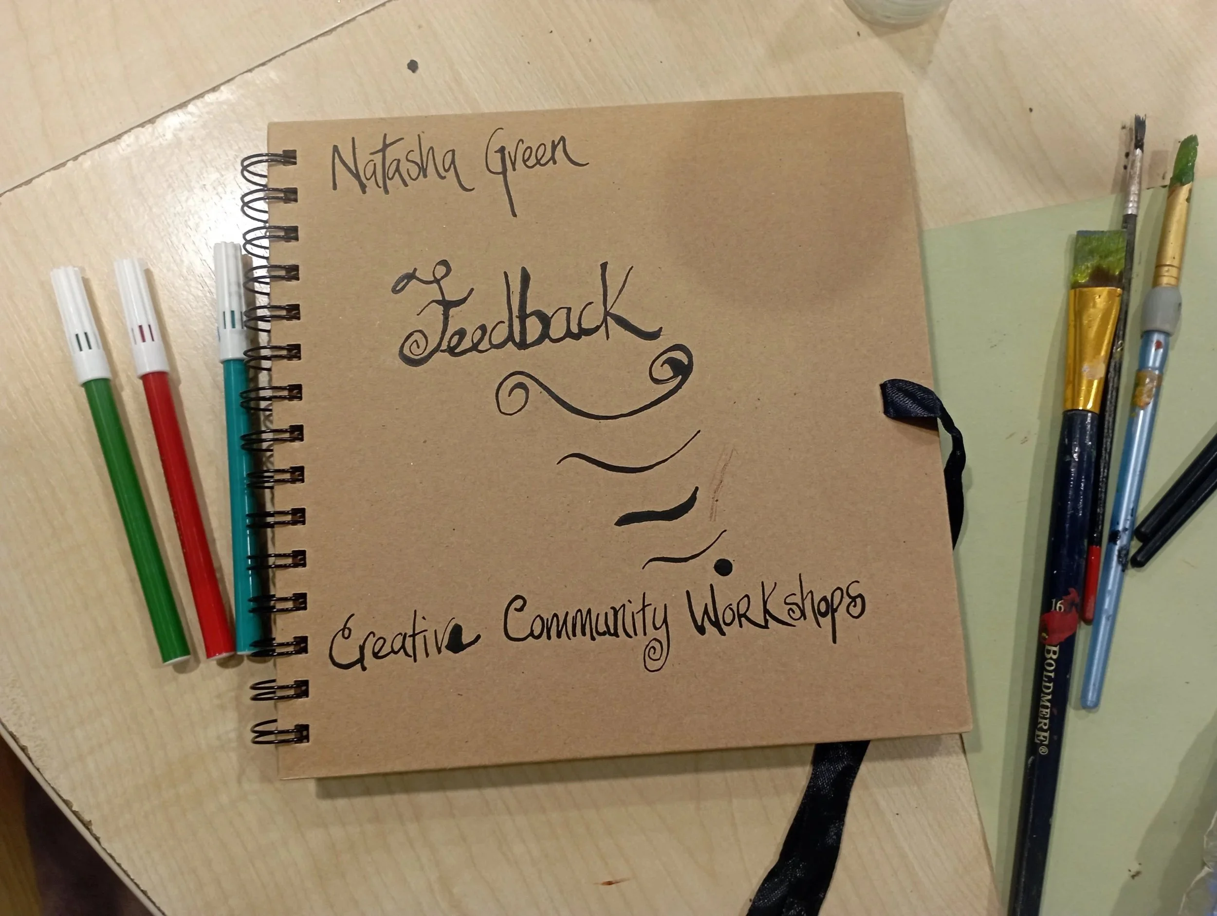 A brown notebook titled 'Feedback' with handwritten text 'Natasha Green' and 'Creative Community Workshops' on the cover, surrounded by colorful markers and paintbrushes on a wooden table.