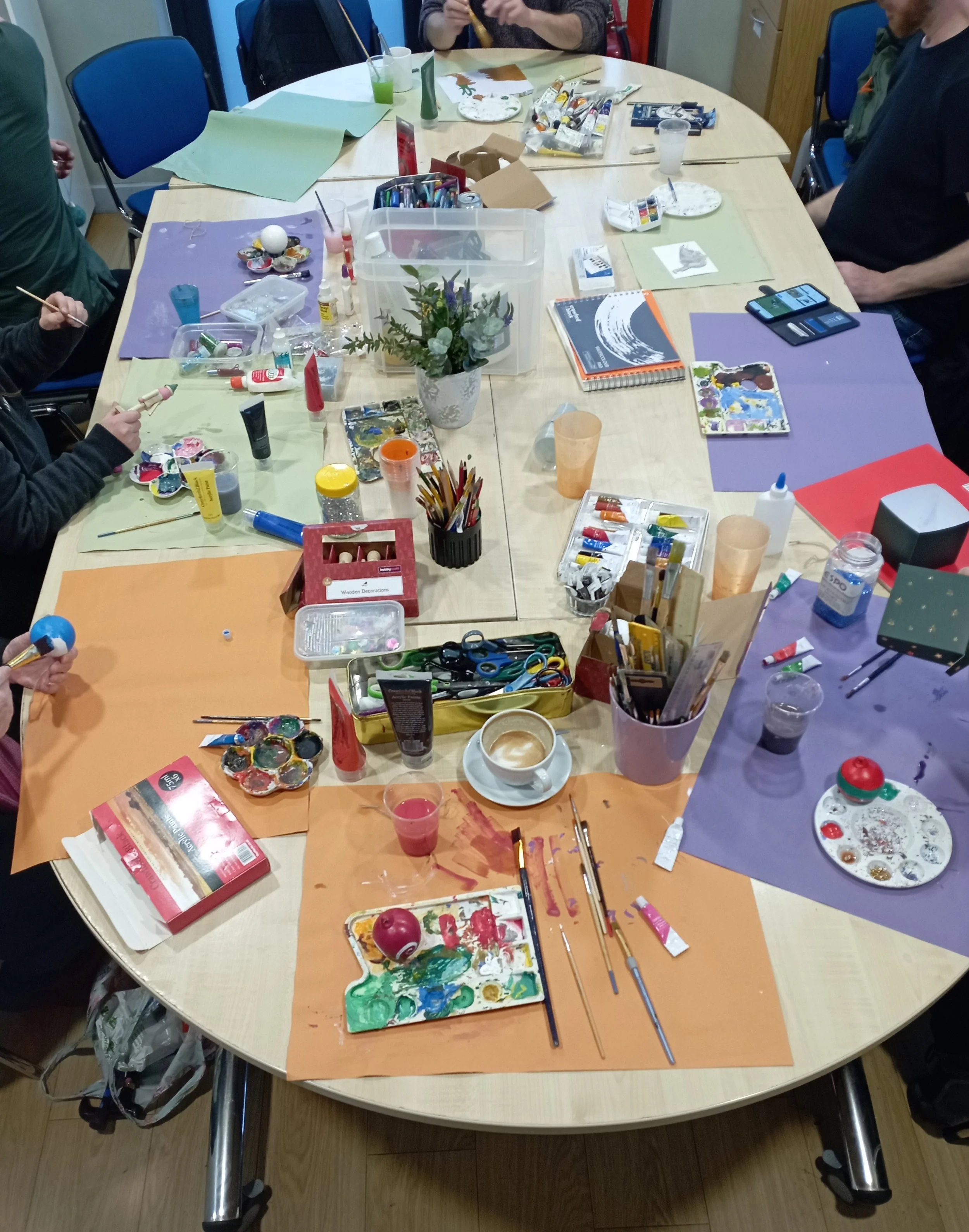A group of people engaged in an arts and crafts activity at a table filled with paint supplies, brushes, paints, scissors, and decorative items, with some drinks and a potted plant in the center.
