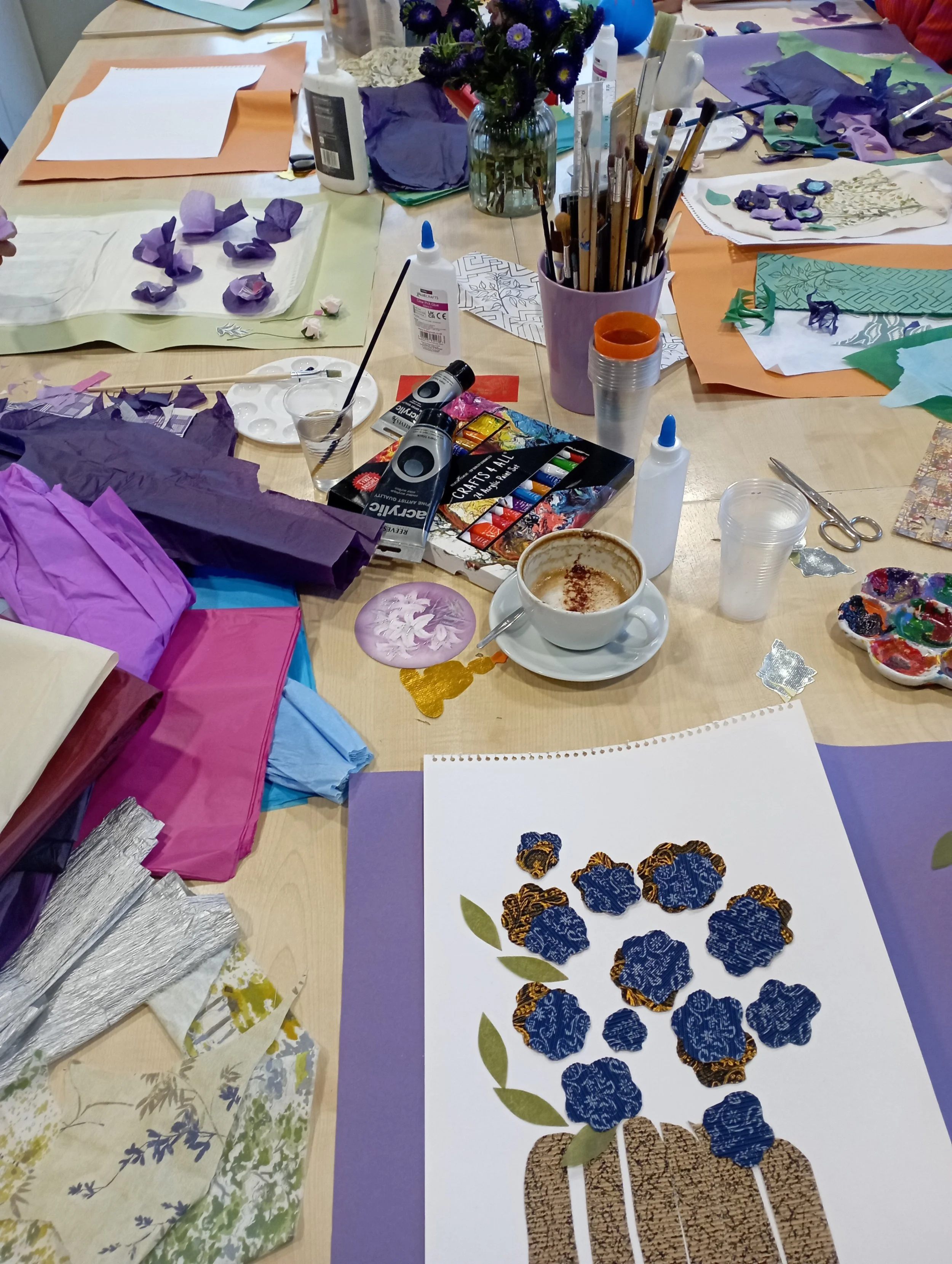 An arts and crafts workspace filled with paper, paint, glue, scissors, and various craft supplies. There is a vase with purple flowers, a coffee cup, and a colorful artwork of a floral arrangement made with fabric or paper flowers.