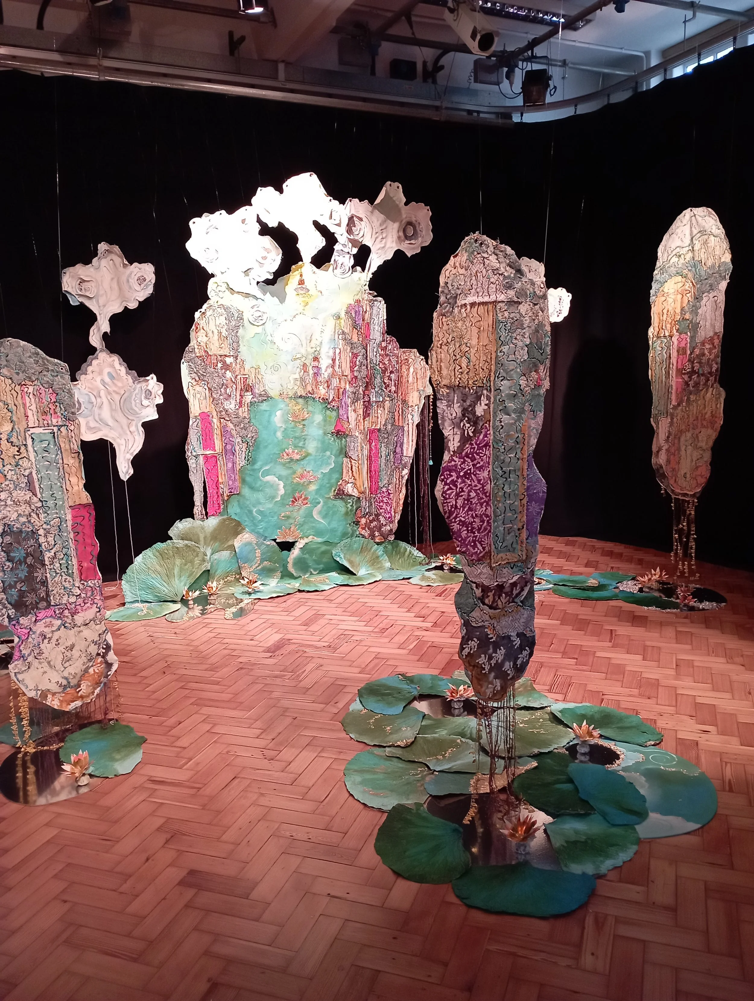 An art installation featuring hanging, fabric-like structures resembling tall rocks or waterfalls, with painted lily pads and water lilies on the floor, set against a black background.