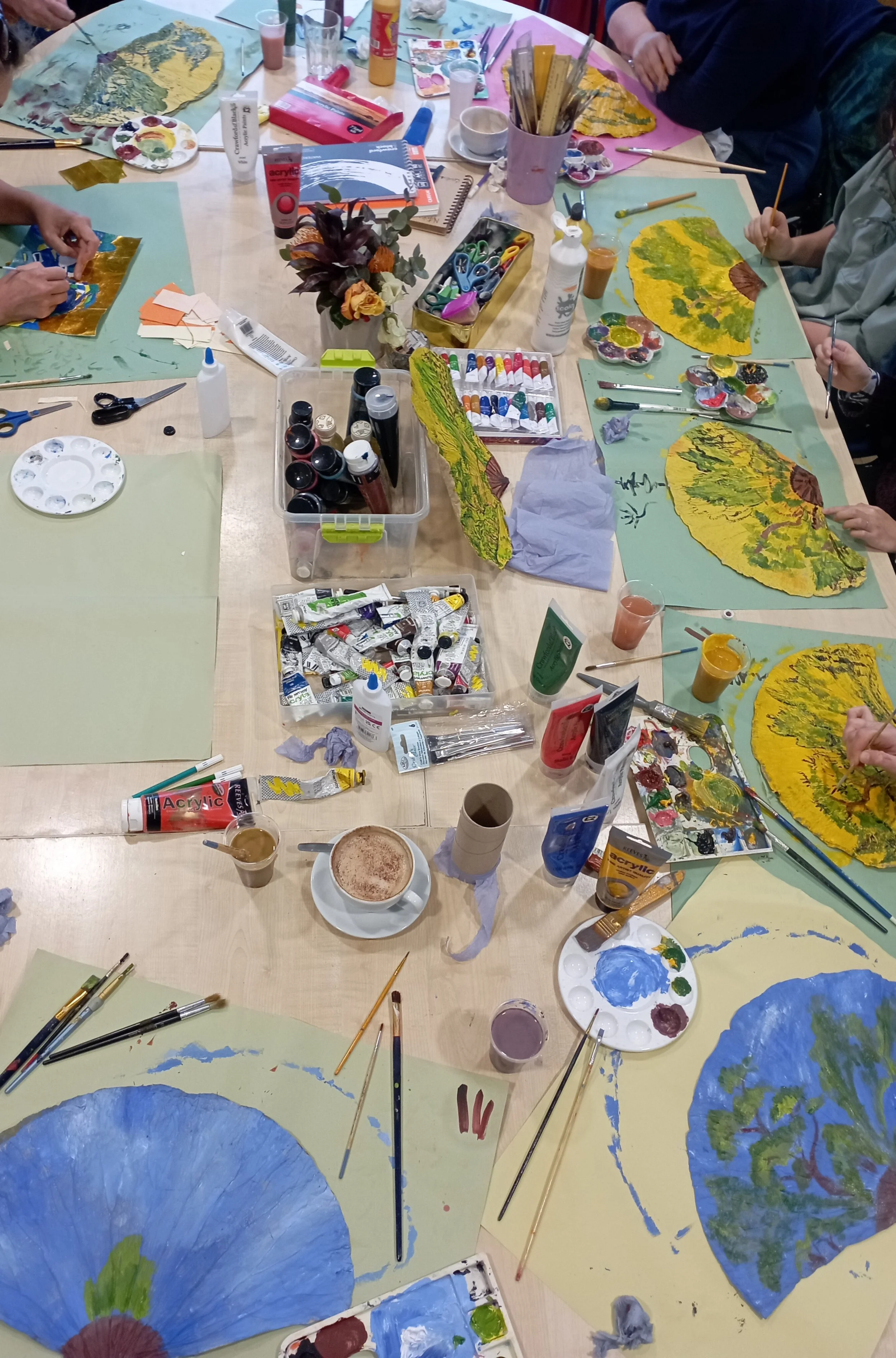 People engaged in a watercolor painting class, creating nature-themed artwork with fans resembling trees and forests, surrounded by art supplies like paints, brushes, and water cups.