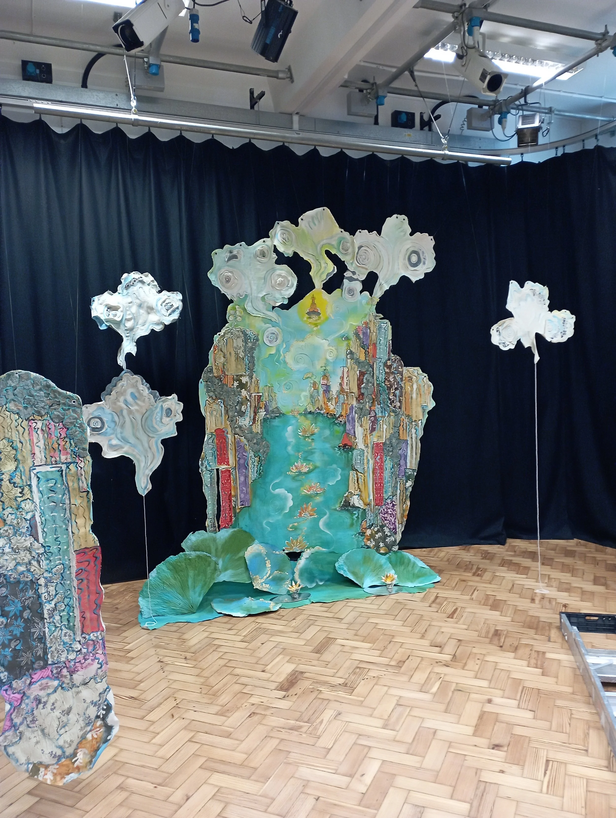 A stage setup inspired by Van Gogh's 'Starry Night,' featuring a large painted backdrop with a river and cityscape, surrounded by paper clouds and lotus flowers, with wooden flooring and stage lighting equipment above.