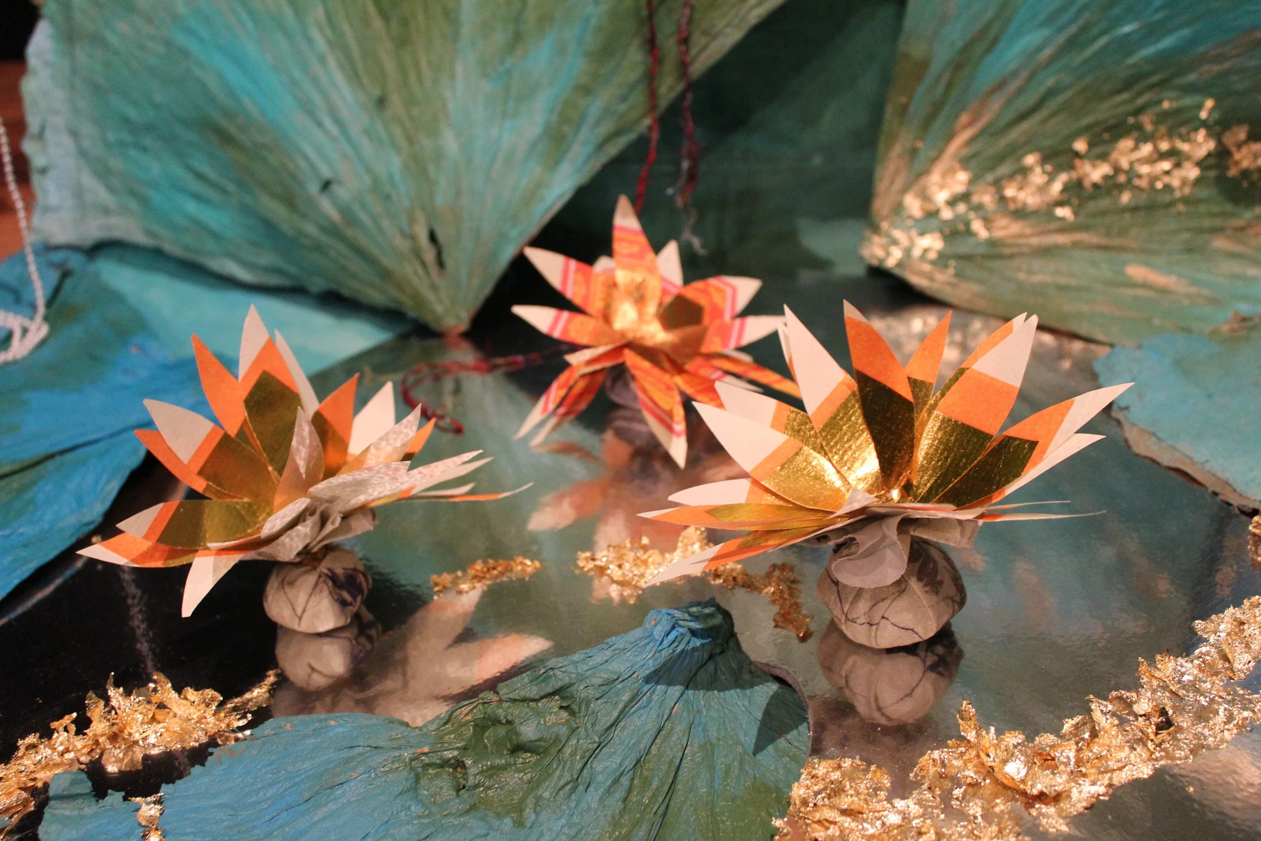 Decorative paper flowers made from gold, orange, and white paper surrounded by painted leaves and gold accents on a reflective surface.