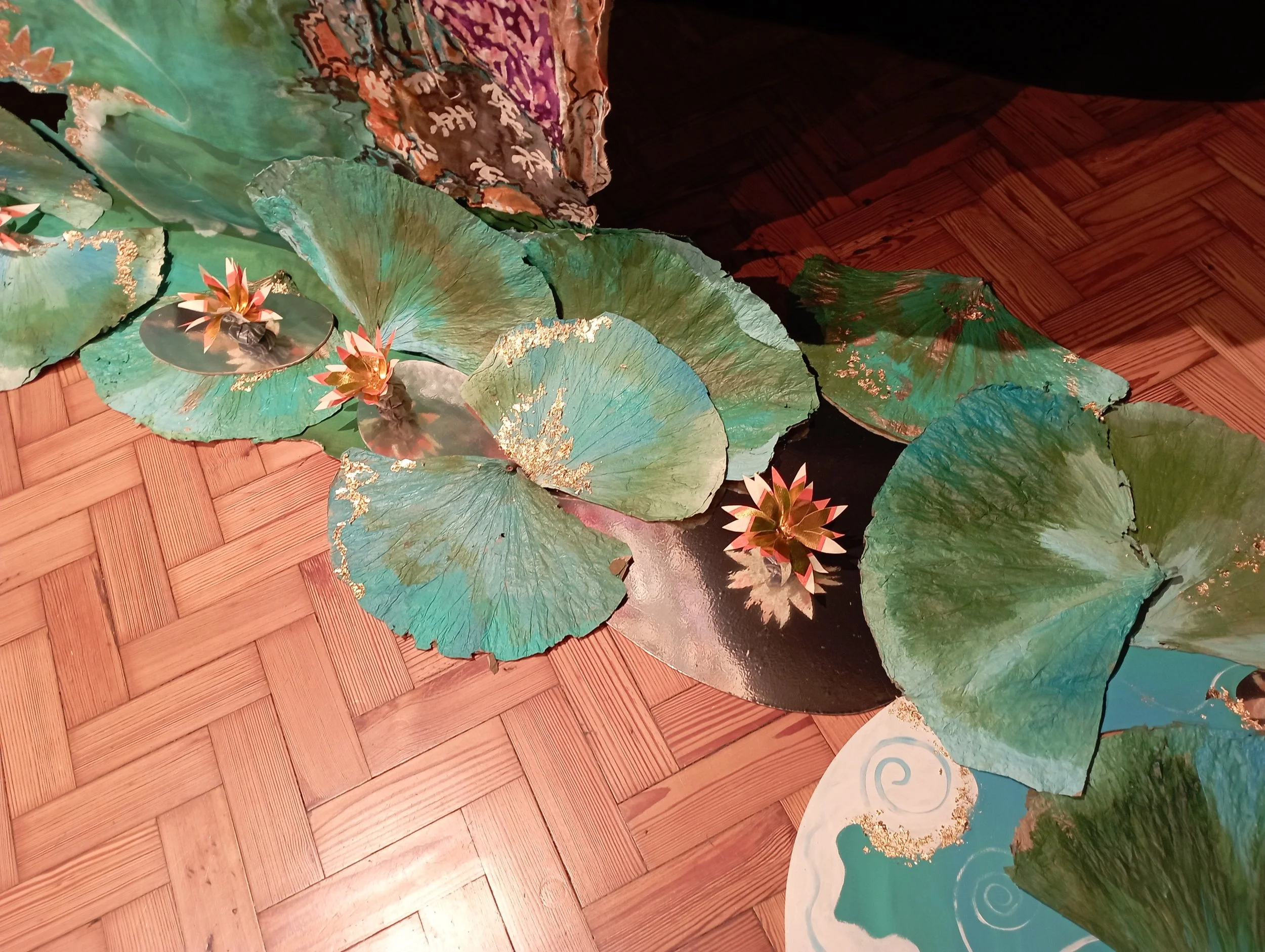 Decorative artificial pond with large green lily pads, pink and orange water lilies, on a wooden floor.