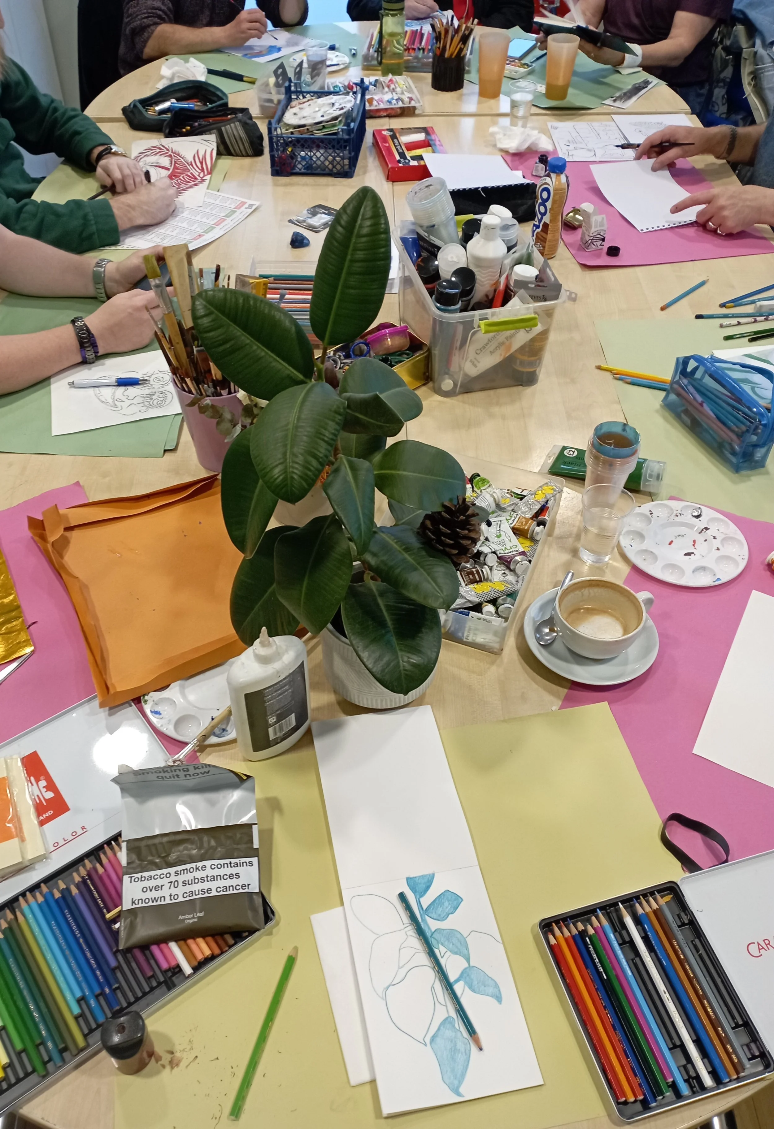 A craft or art workshop with various colored papers, colored pencils, paint supplies, and a potted plant on a table. People are seated around the table working on creative projects.