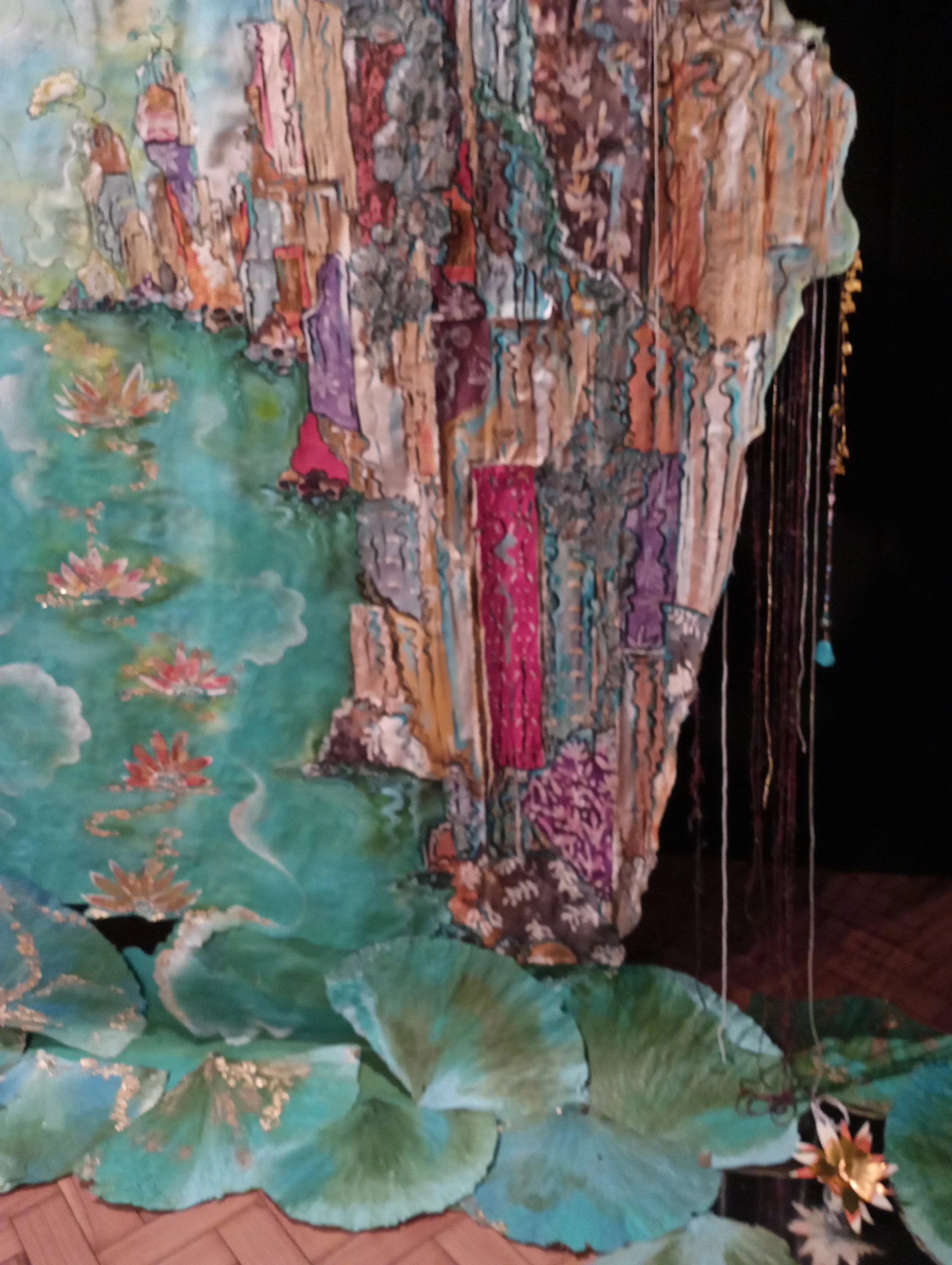 Colorful textile artwork with a landscape scene featuring water lilies, lily pads, and a mountain range with detailed textures and vibrant colors, hanging against a dark background.