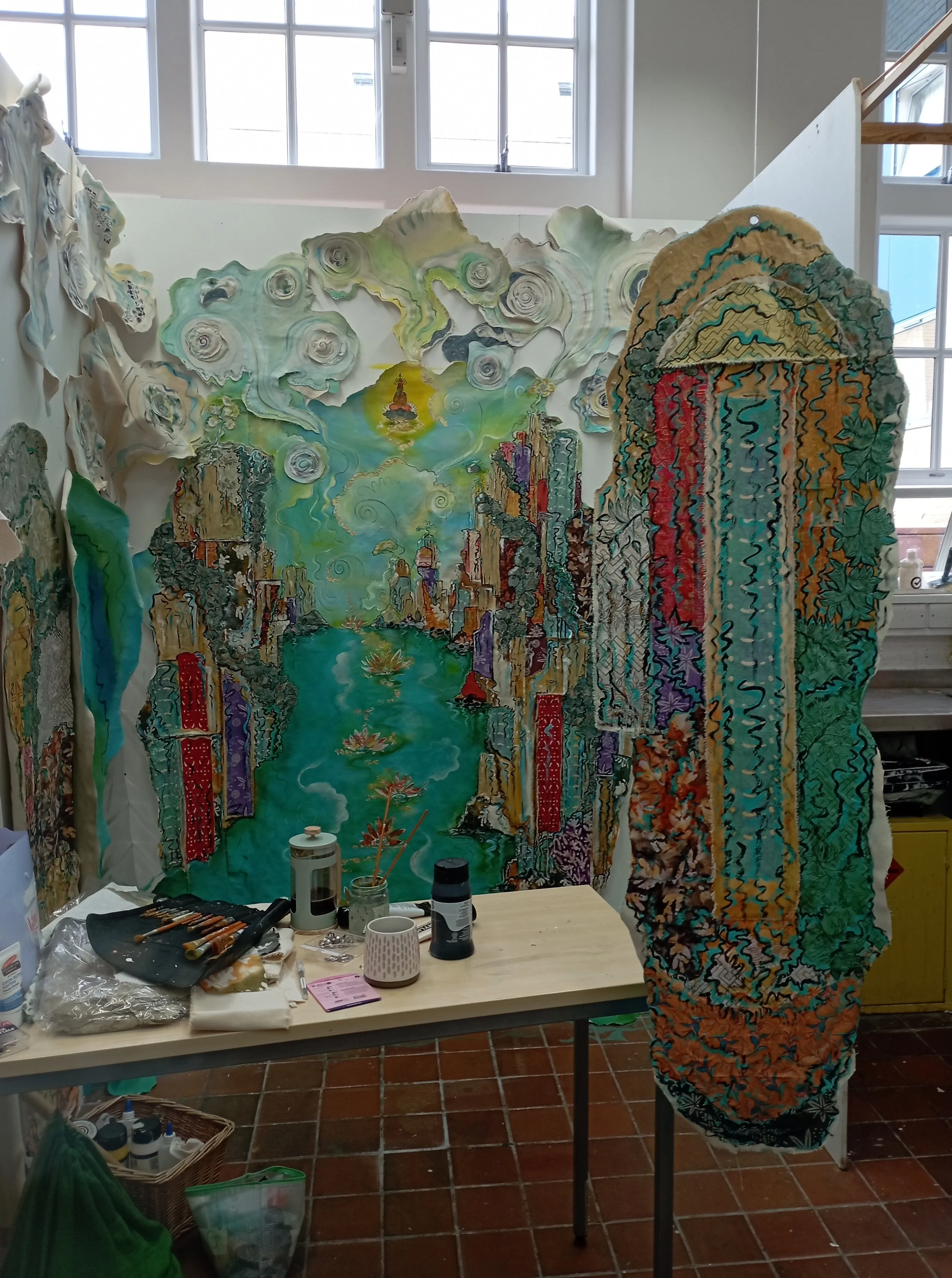 A colorful, textured artwork resembling a cityscape and river scene, with a cloudy sky and a glowing sun above in an indoor art studio.