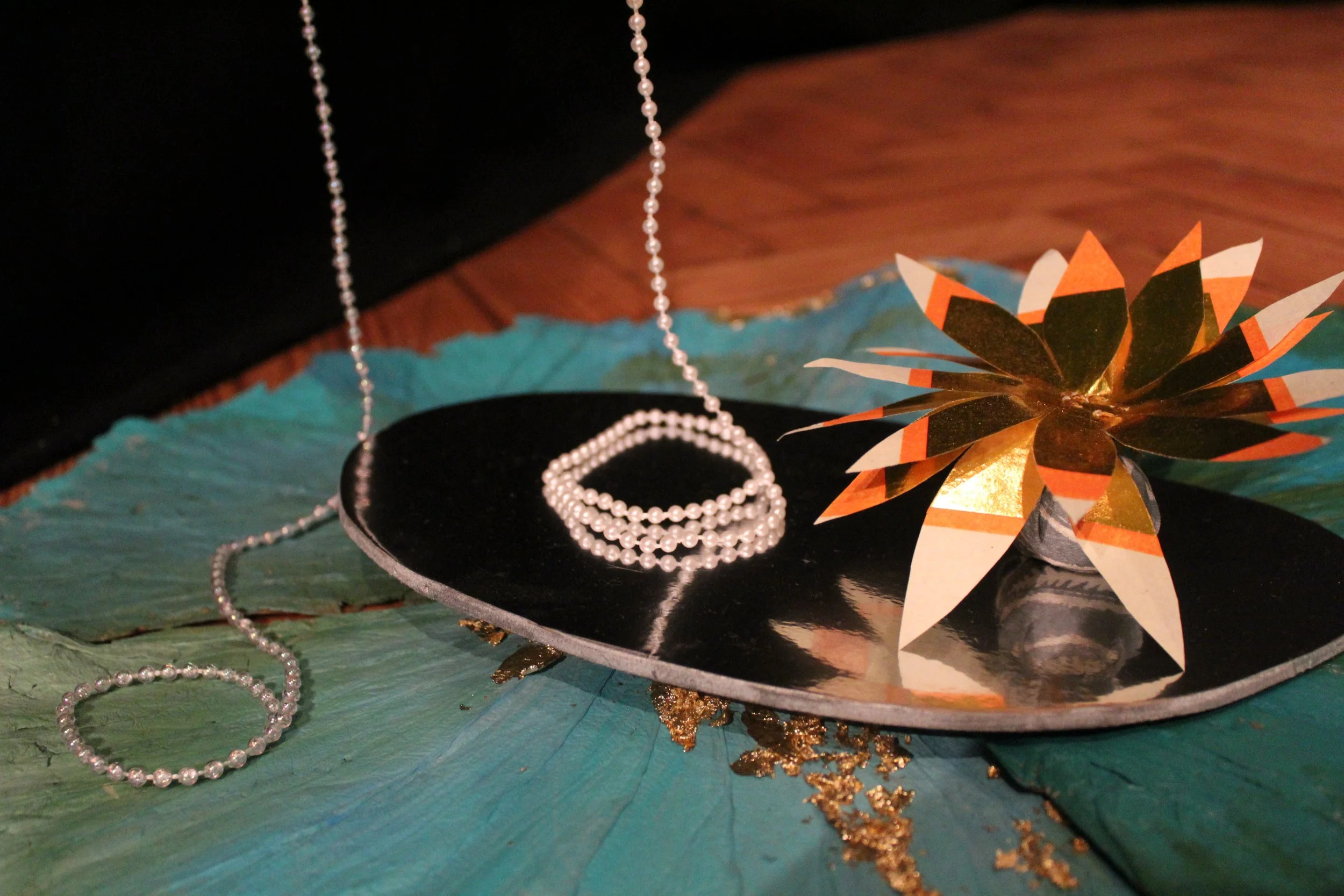 Pearl jewelry, including a necklace, bracelet, and dangling chain, displayed on a black circular tray set on a textured, colorful surface with a decorative paper flower with orange, black, white, and gold accents nearby.