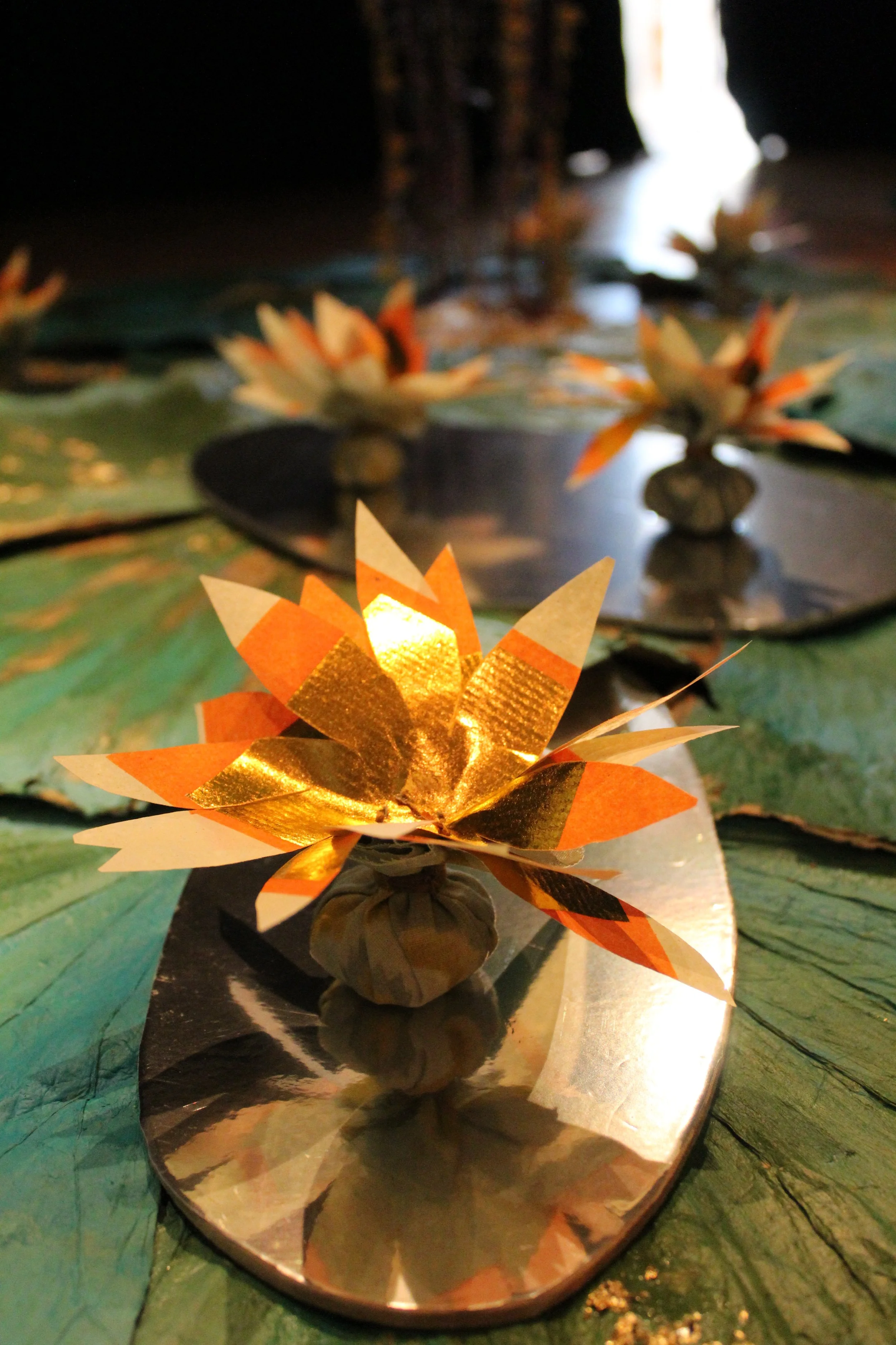 Close-up of decorative gold and orange paper flowers placed on reflective circular surfaces, with additional flowers and greenery in the background, creating an elegant arrangement.