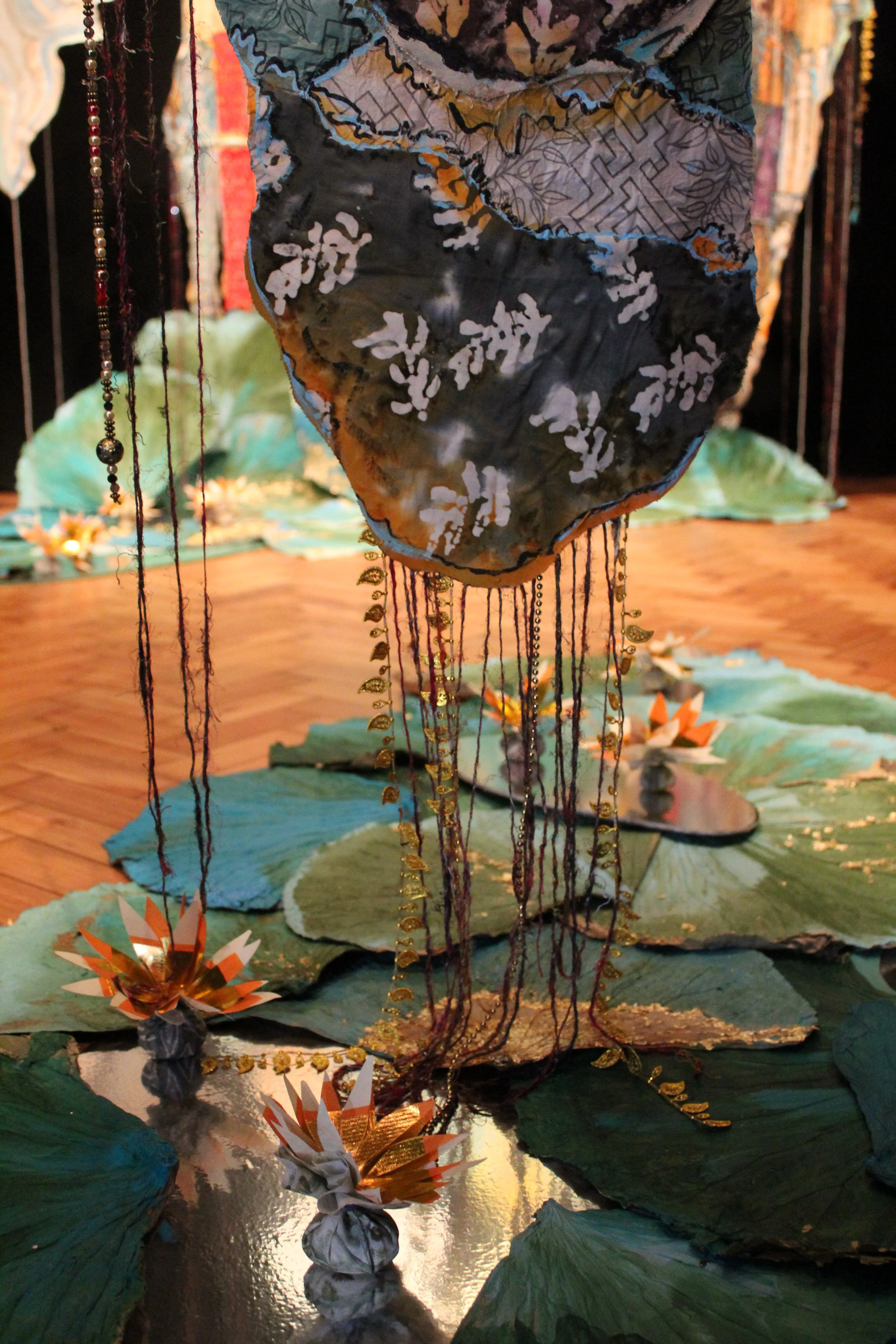 An art installation featuring a hanging fabric sculpture resembling a flower or cloud, with hanging beads and chains, surrounded by paper flowers and large paper lily pads on a reflective surface, in a gallery setting.