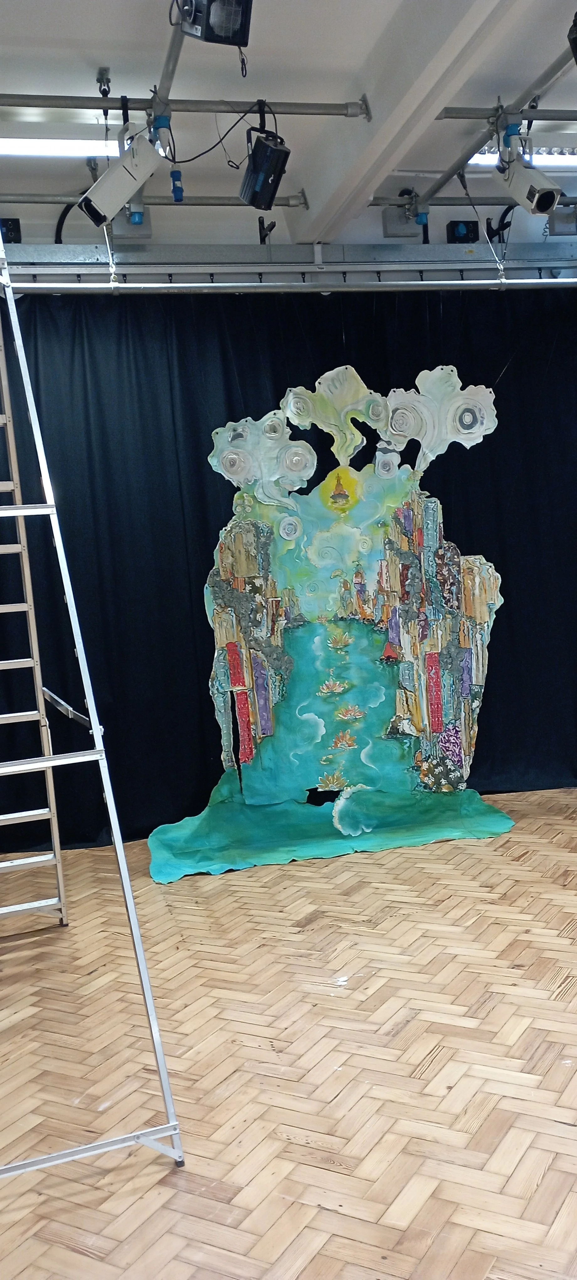 Set piece with painted scenery of a river flowing between cliffs and lush greenery, with floating water lilies. The piece is shaped like a large, colorful human figure with clouds above its head, and is set against a black curtain in a studio space w