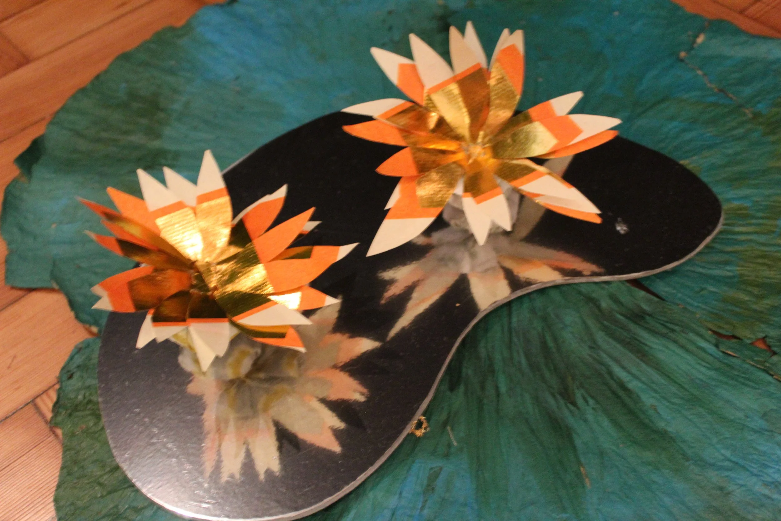 Crafted paper lotus flowers with orange, white, and gold colors placed on a black mirror-like surface, surrounded by large green lotus leaves.