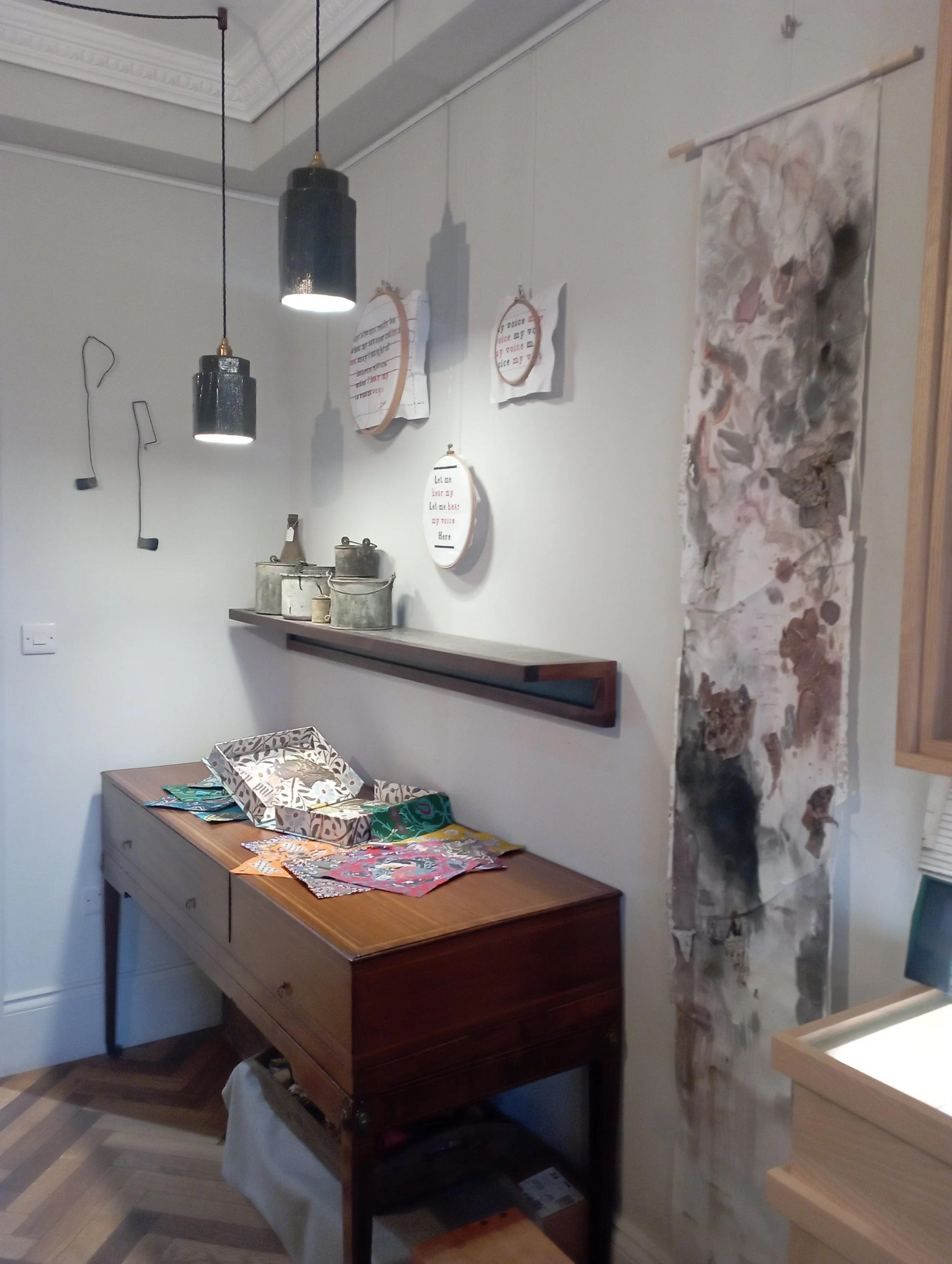 Interior corner of a room with a wooden sideboard, wall decorations including embroidery hoops with text, hanging wall art, and ceiling lights.