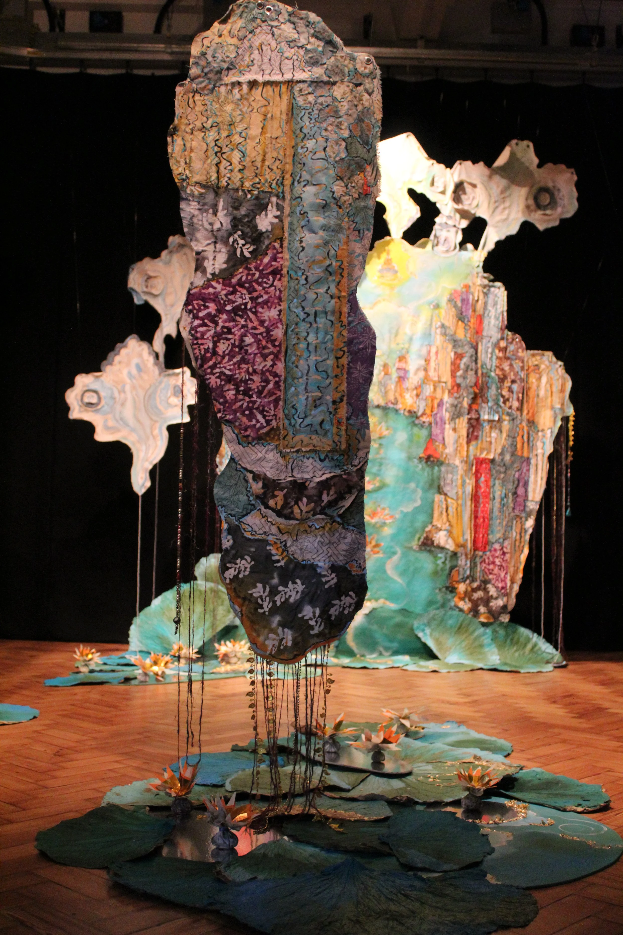 A textile art installation featuring layered fabric pieces hanging and arranged to resemble a floating face or figure, with a backdrop of painted cityscape and water scene, all set on a wooden floor with lily pad shapes and flowers.