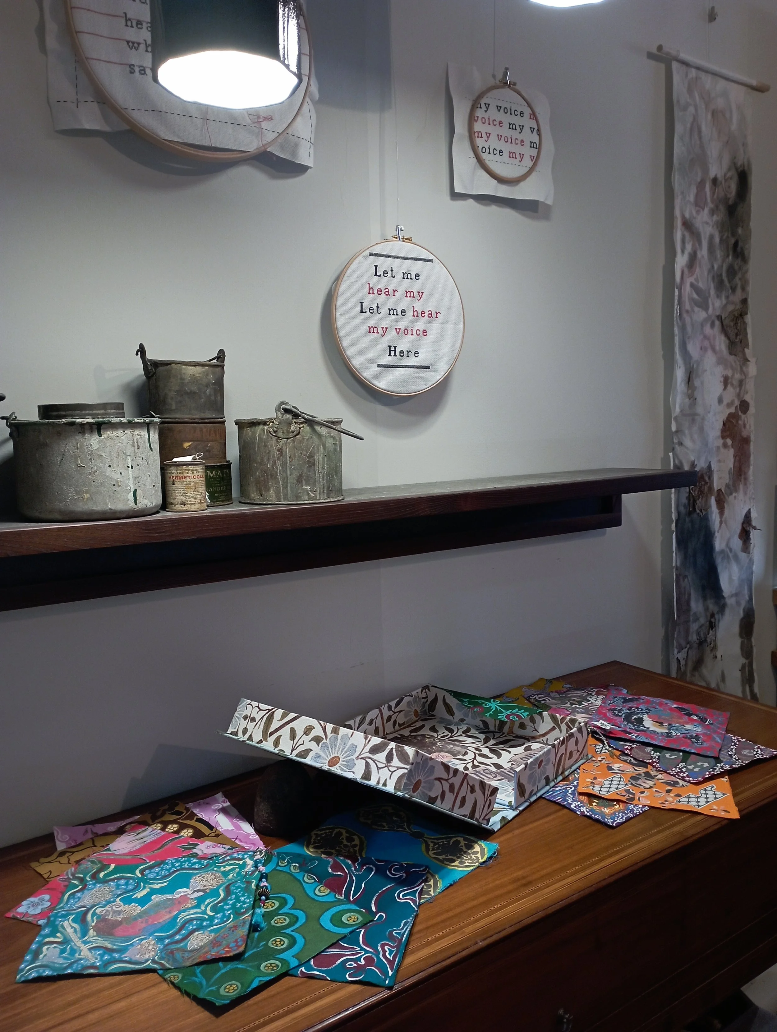 A display of embroidered hoops on a wall, a wooden table with fabric wraps, and vintage metal containers on a shelf.