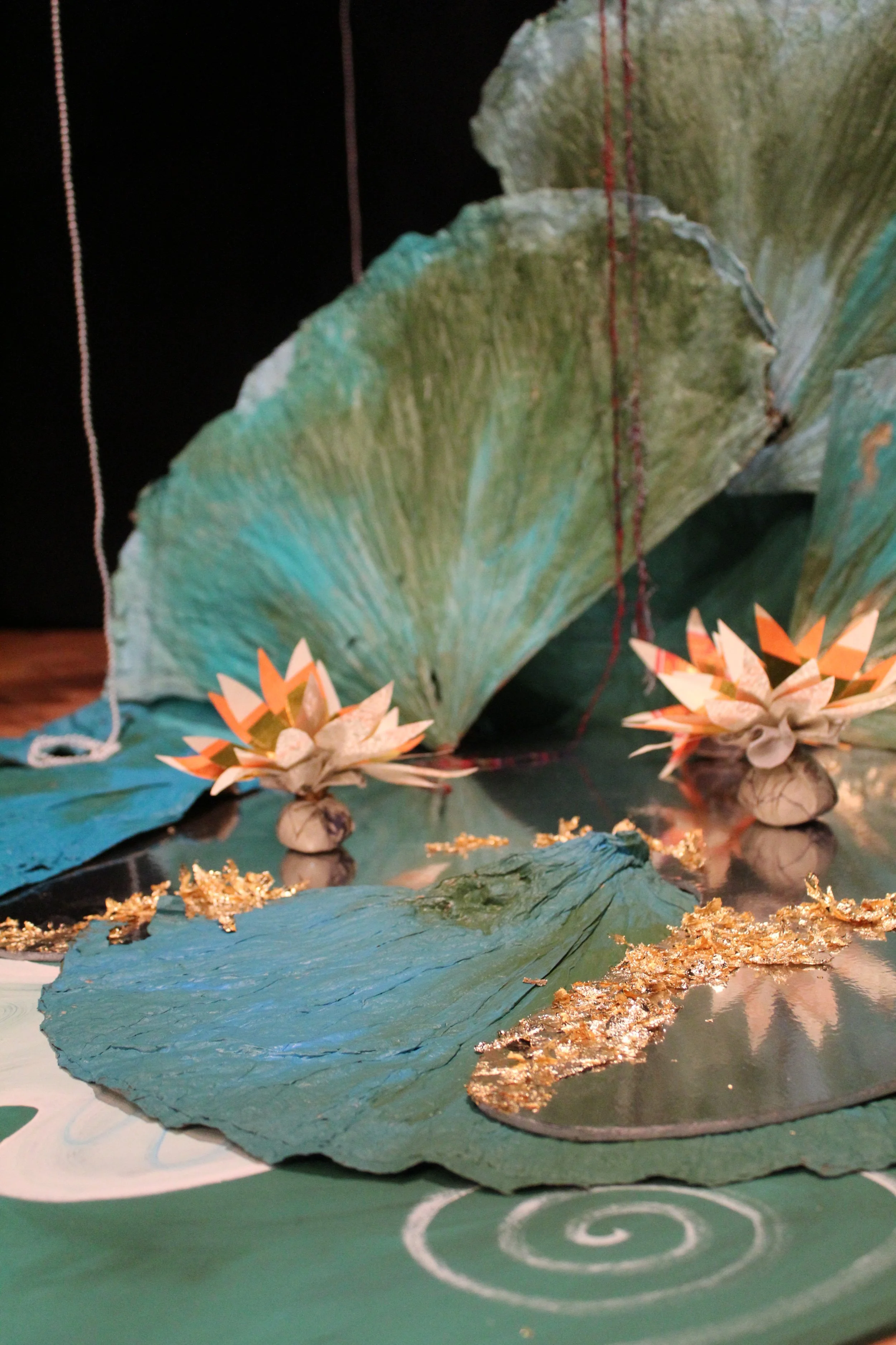 Close-up of a craft project featuring large blue and green paper leaves, paper flowers resembling lilies with orange and cream petals, and gold-painted leaves, all laid on a table with a dark background.