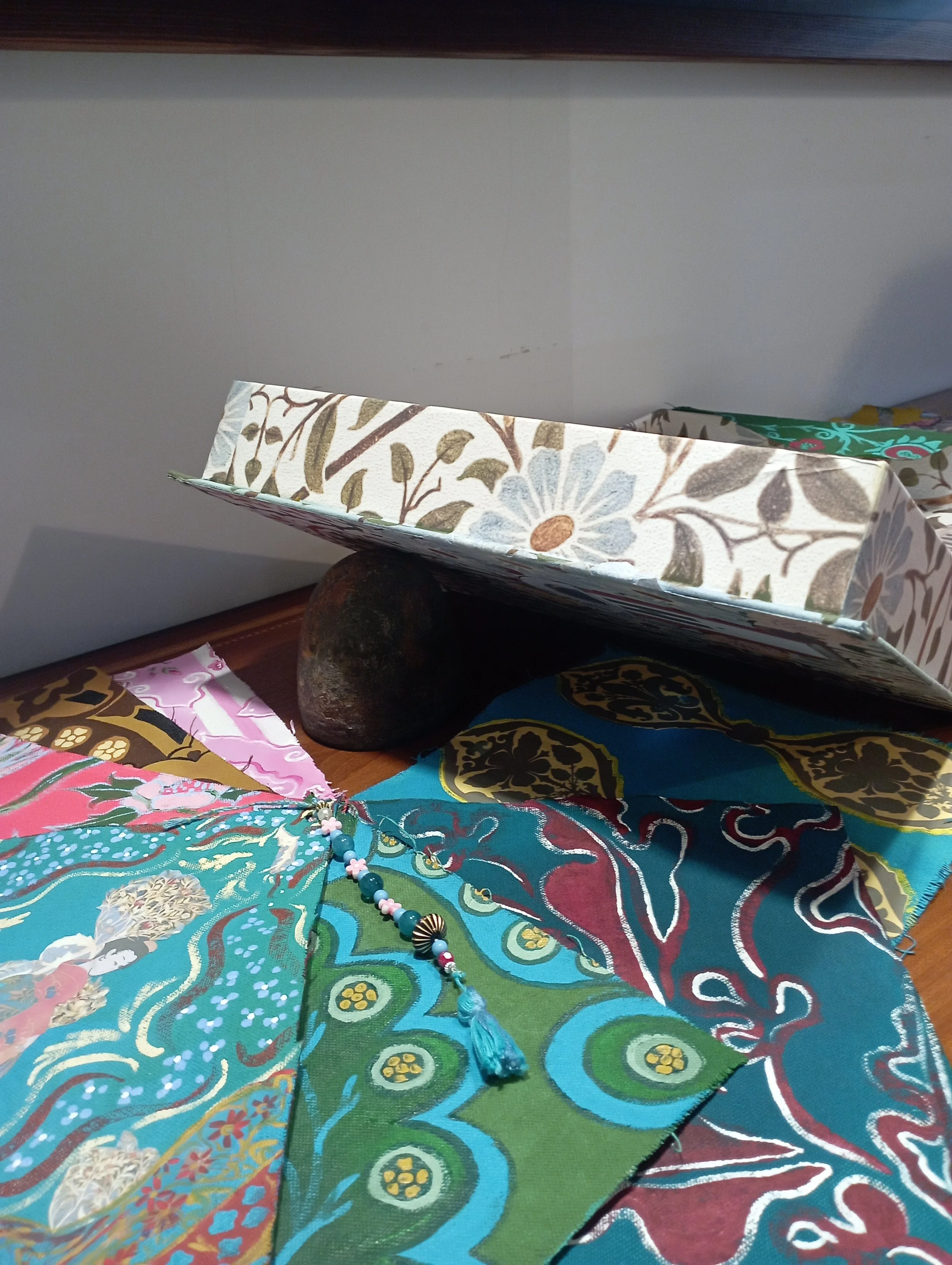 Decorative fabric with multicolored floral and paisley print, some jewelry with beads and tassels, a painted box on a wooden surface, supported by a stone, against a plain white wall.