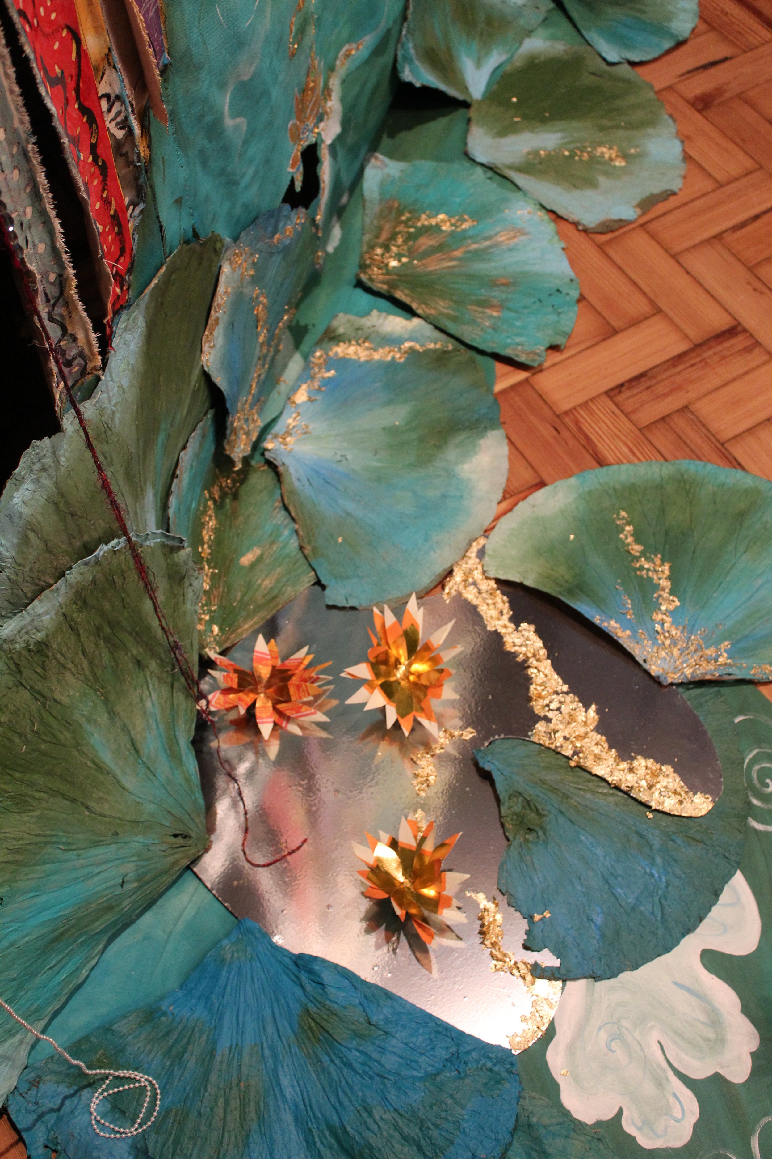Decorative scene with large green lily pads, three ornate paper flowers with gold accents, and a reflective silver surface, set against a patterned green background.