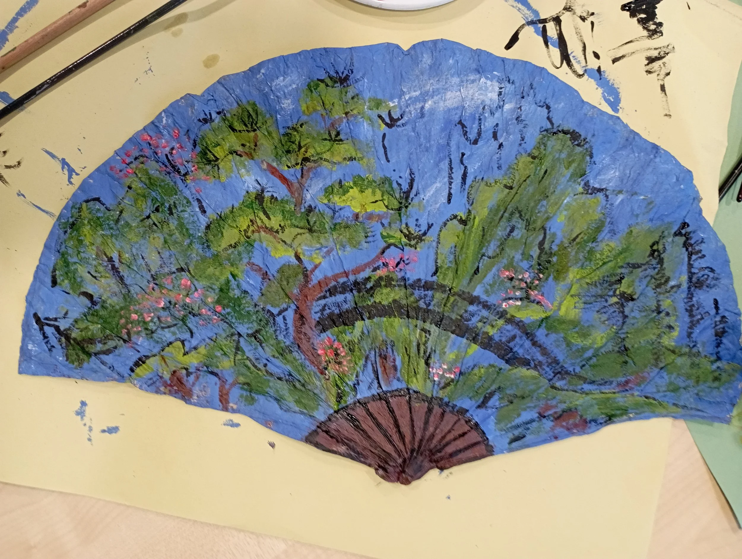 Decorative hand-painted paper fan featuring trees, foliage, and blossoms on a blue background.