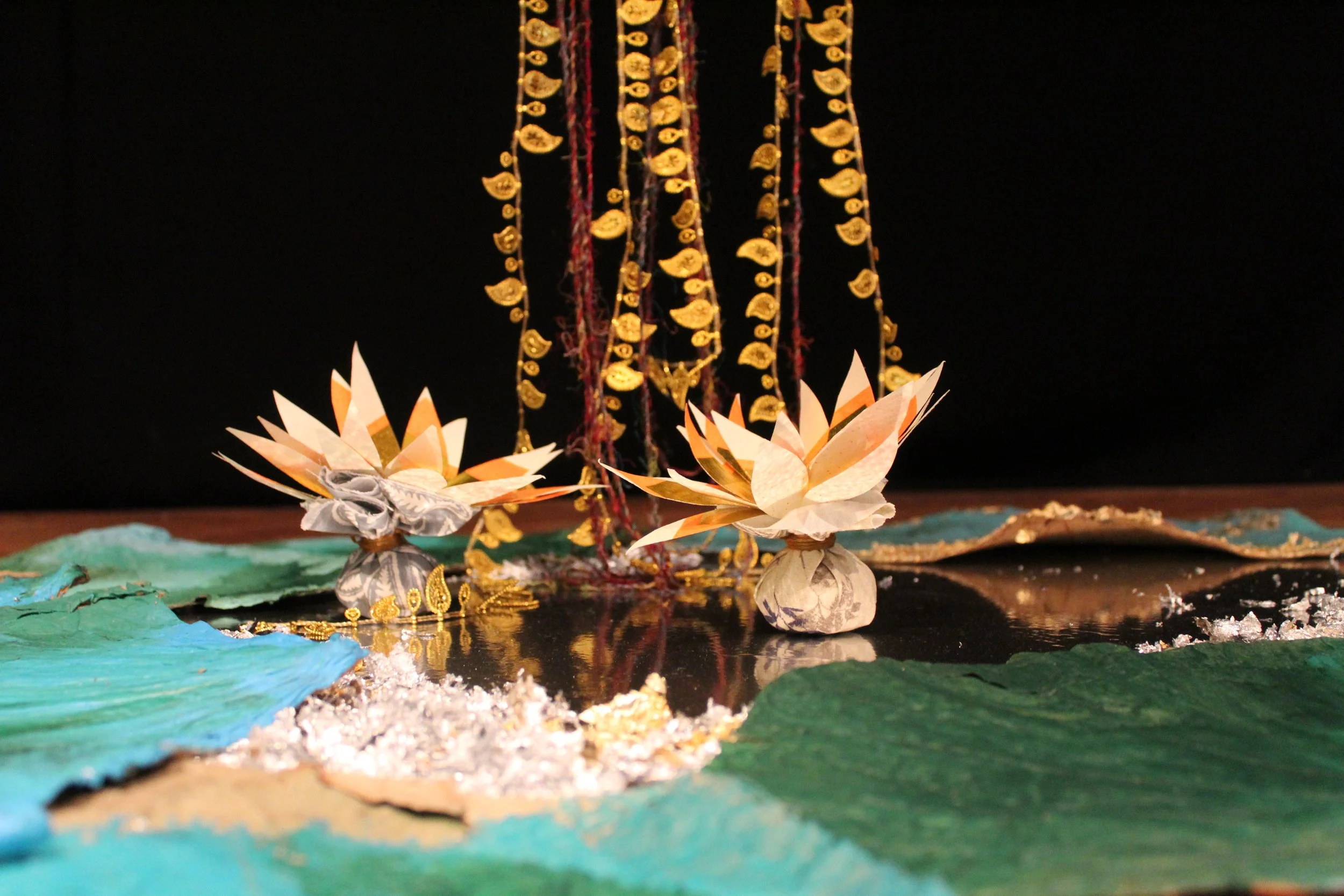 Decorative scene with two paper lotus flowers, colorful paper leaves in green and blue, and hanging gold beads against a black background.