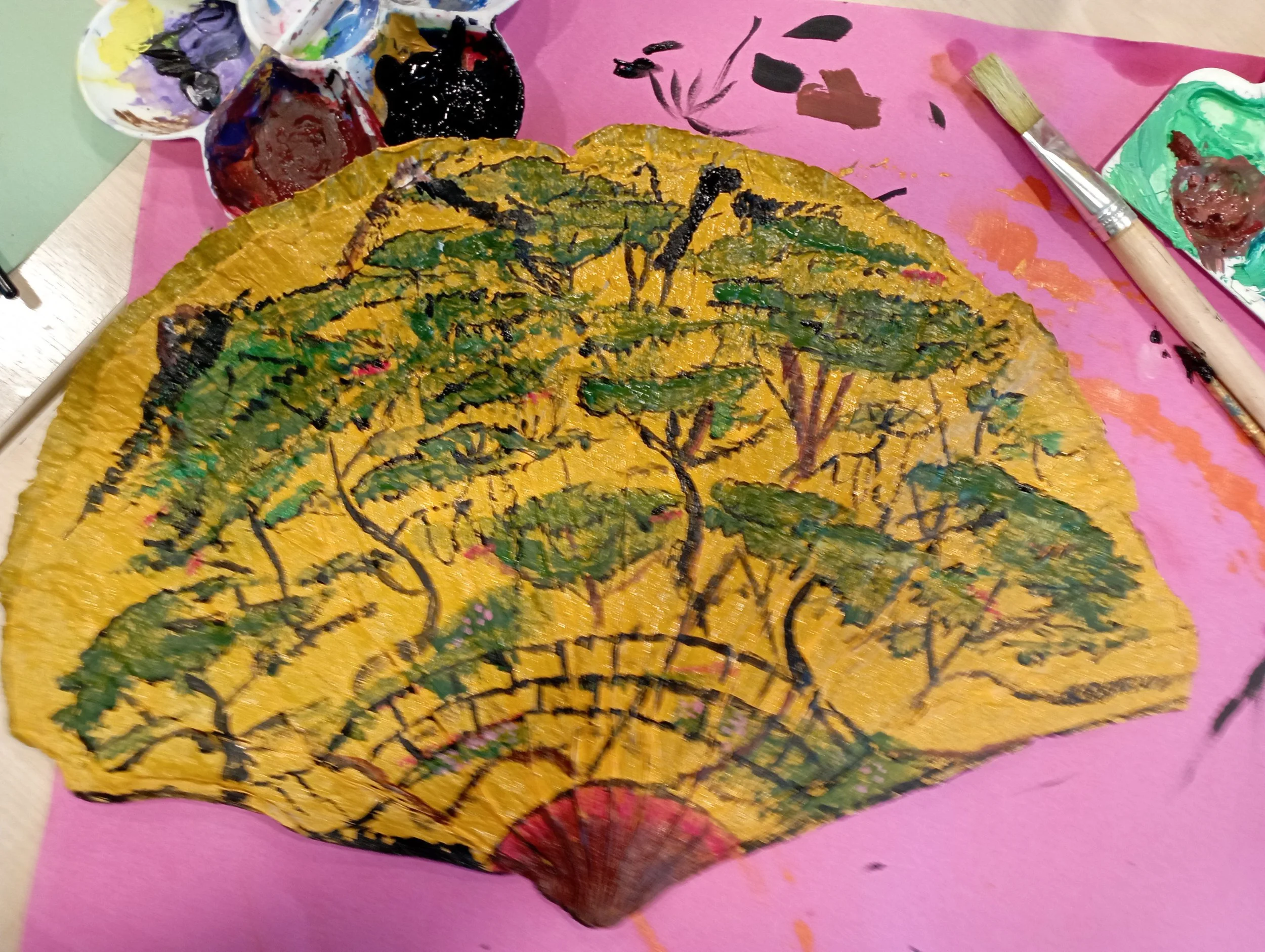 A painted decorative hand fan with a landscape scene of trees, on yellow background. Art supplies, including paint, a palette, and brushes, are visible around it.