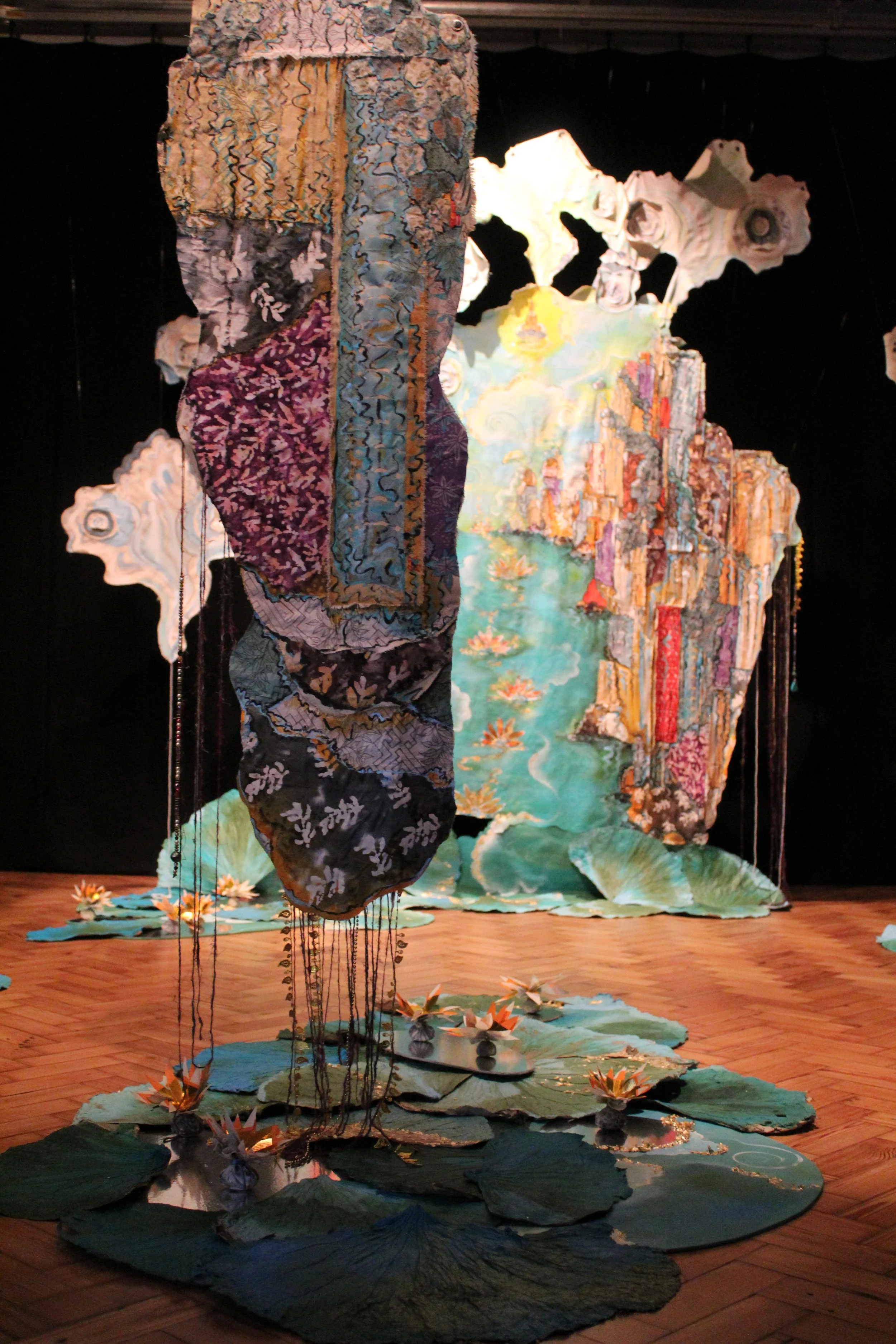 An intricate art installation features layered fabrics, joss paper, acrylic and canvas with mirror pools with floating lotus flowers and dried and painted lotus leaves. Masters Project 2025 by Natasha Green.