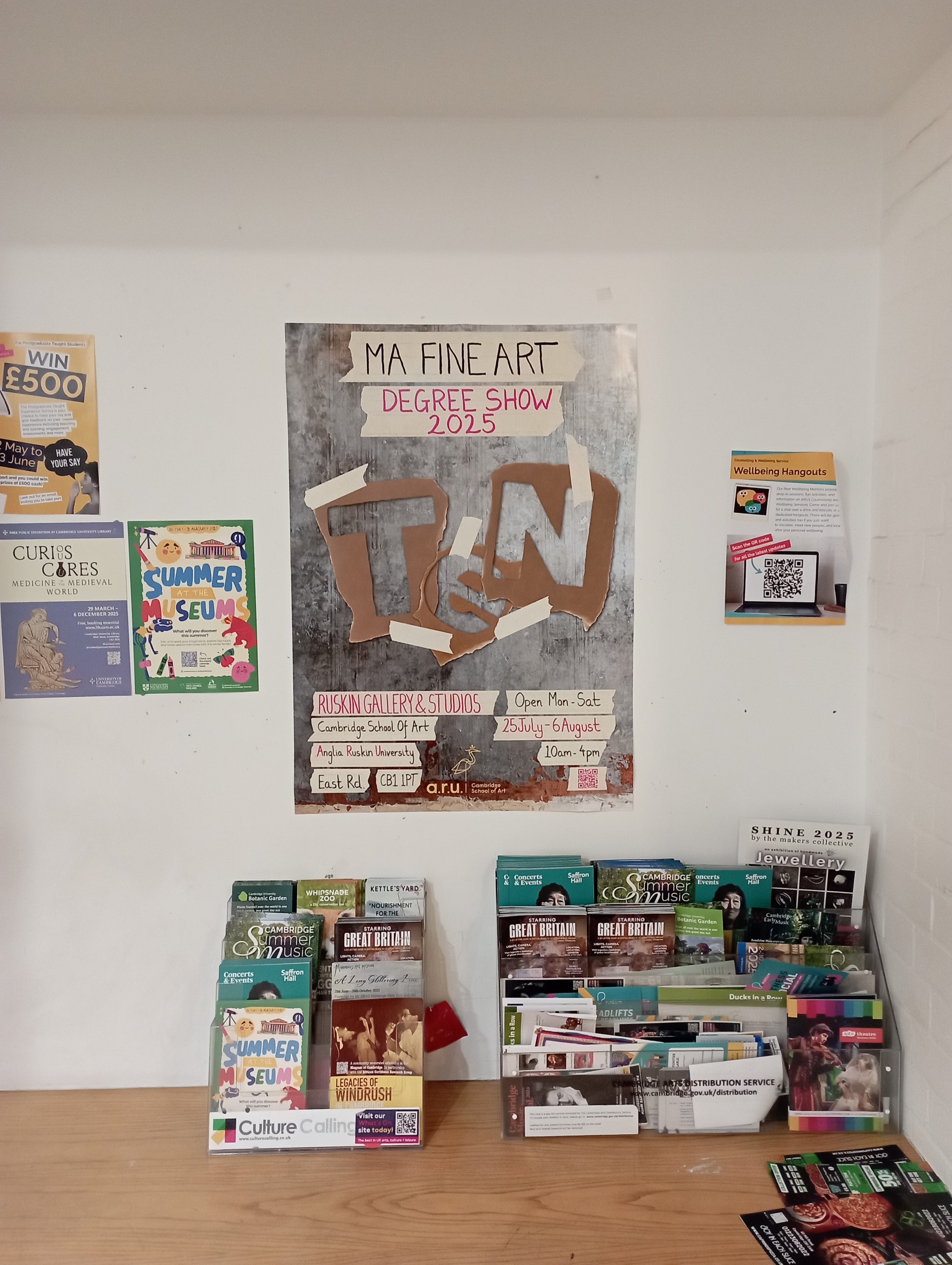 Poster advertisement for MA Fine Art Degree Show 2025 at Ruskin Gallery & Studios, Cambridge School of Art, showing event dates and details on a white wall, with brochure and flyer displays on a wooden table below.