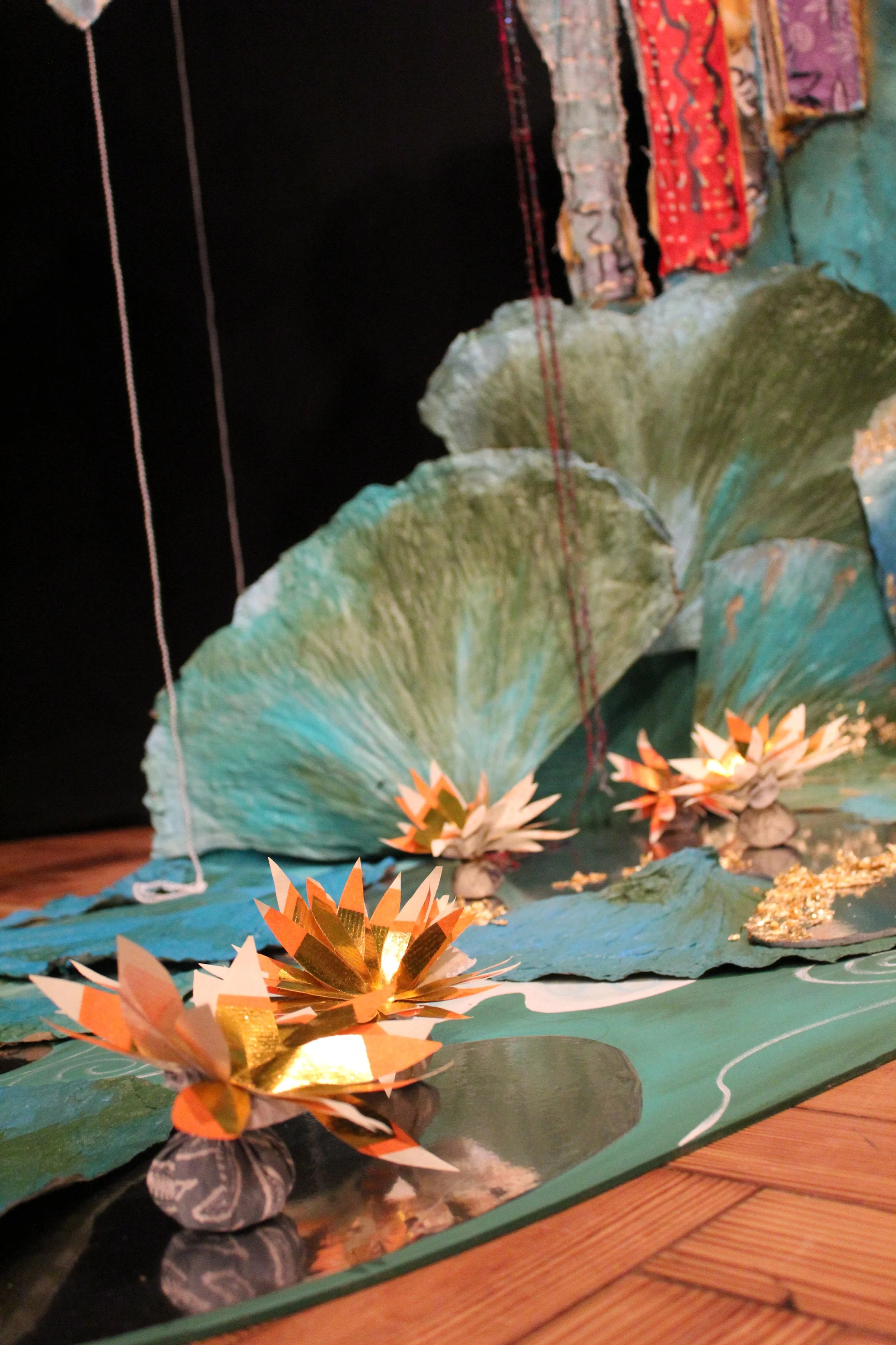 Art installation of paper flowers and large leaf shapes, with string hanging from the top, set against a black background.