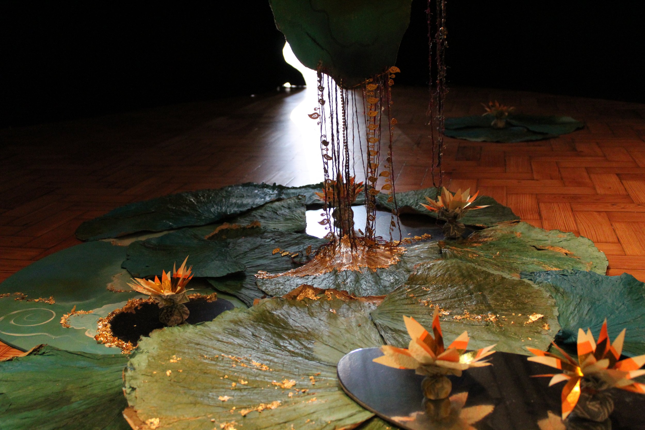 An art installation with large green lily pads, paper lotus flowers, hanging decorative chains, and a reflective mirror on a wooden floor, with a dark background and a bright light source in the distance.