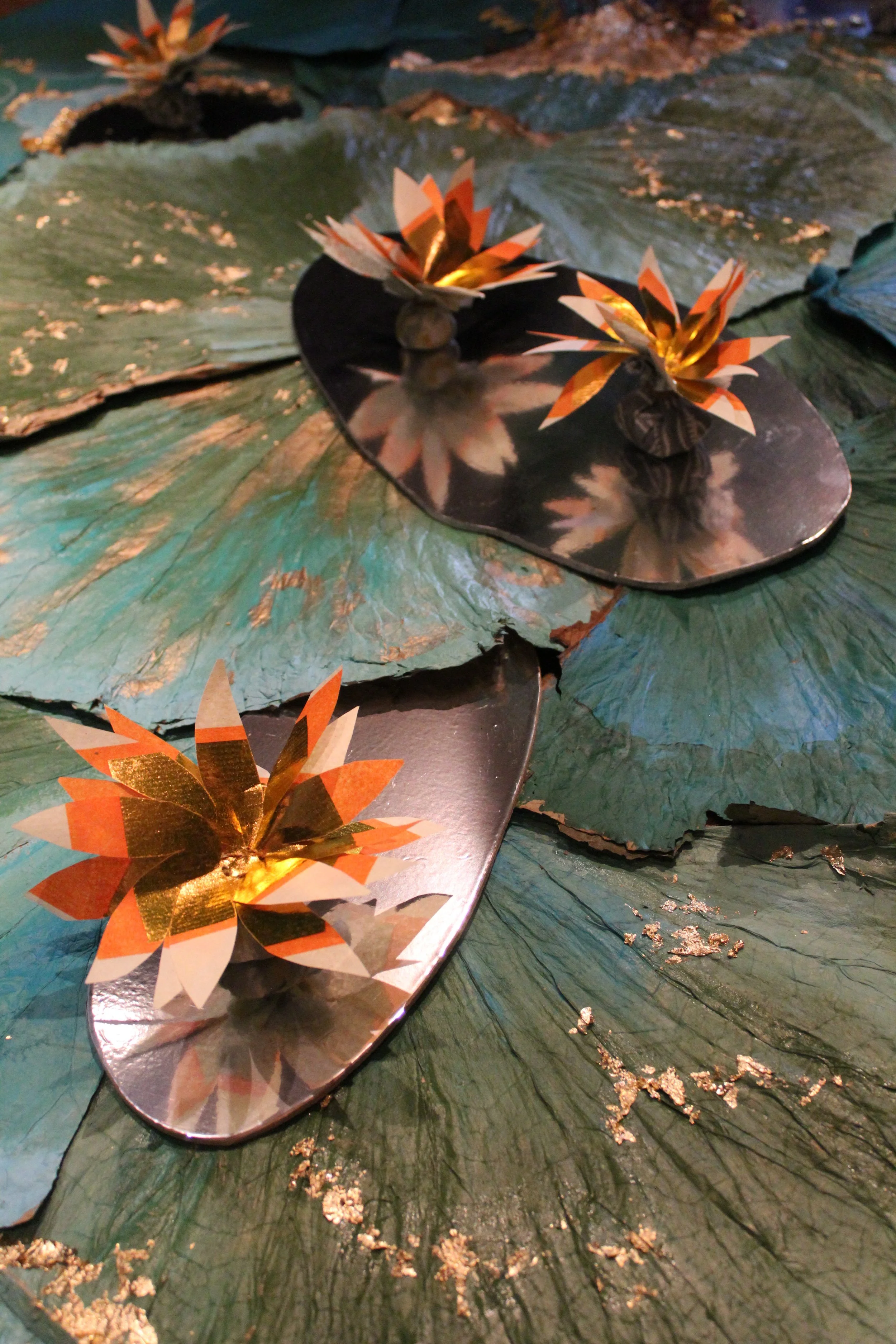 Close-up of a decorative water lily arrangement with metallic gold and orange paper flowers floating on green lily pads.