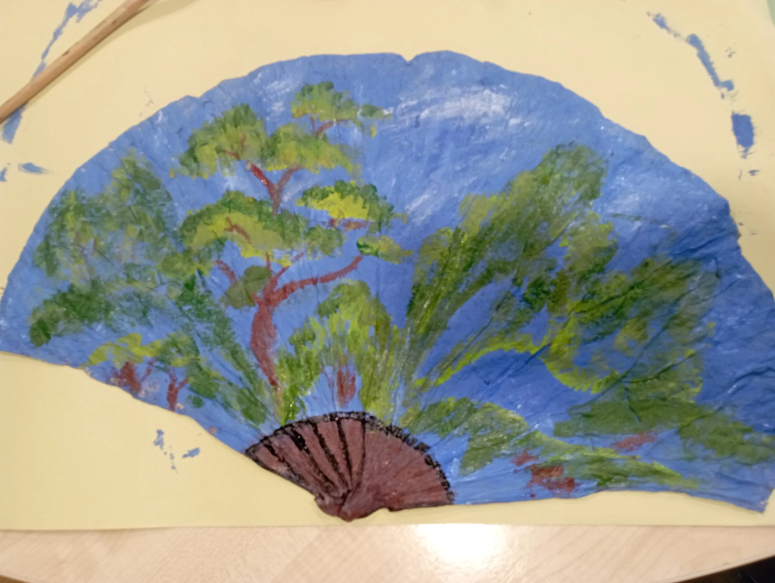 Hand-painted blue paper fan with green trees and brown branches and trunk, on yellow background.