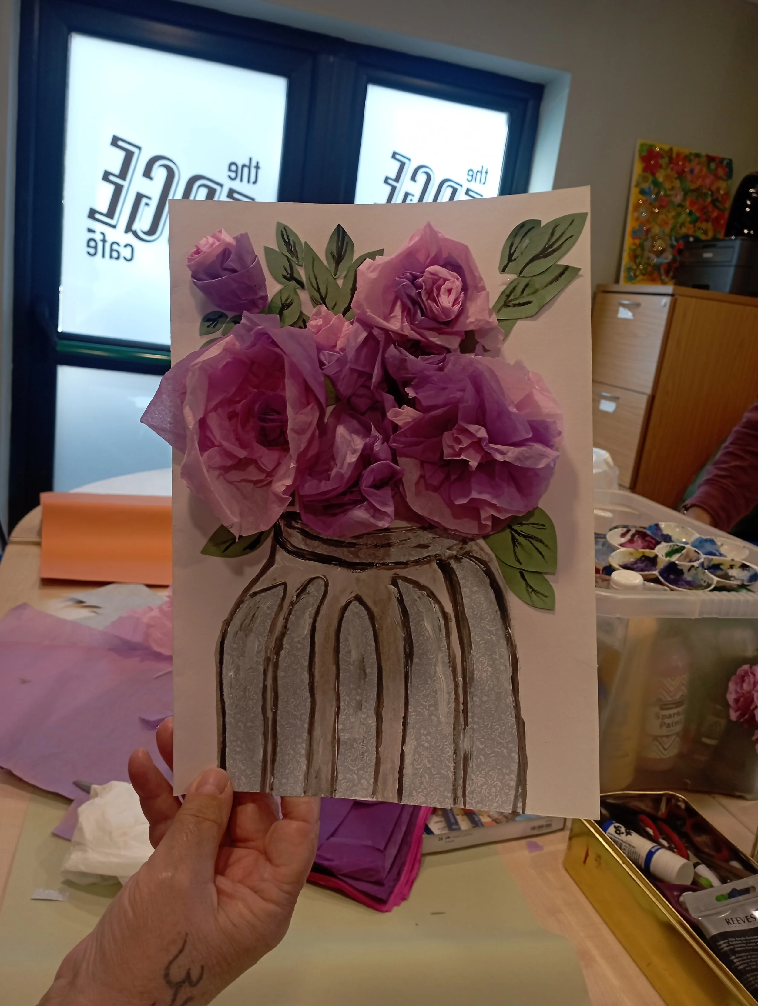 A person holding an art piece with a painted vase and pink tissue paper flowers. The background shows a room with windows and art supplies.