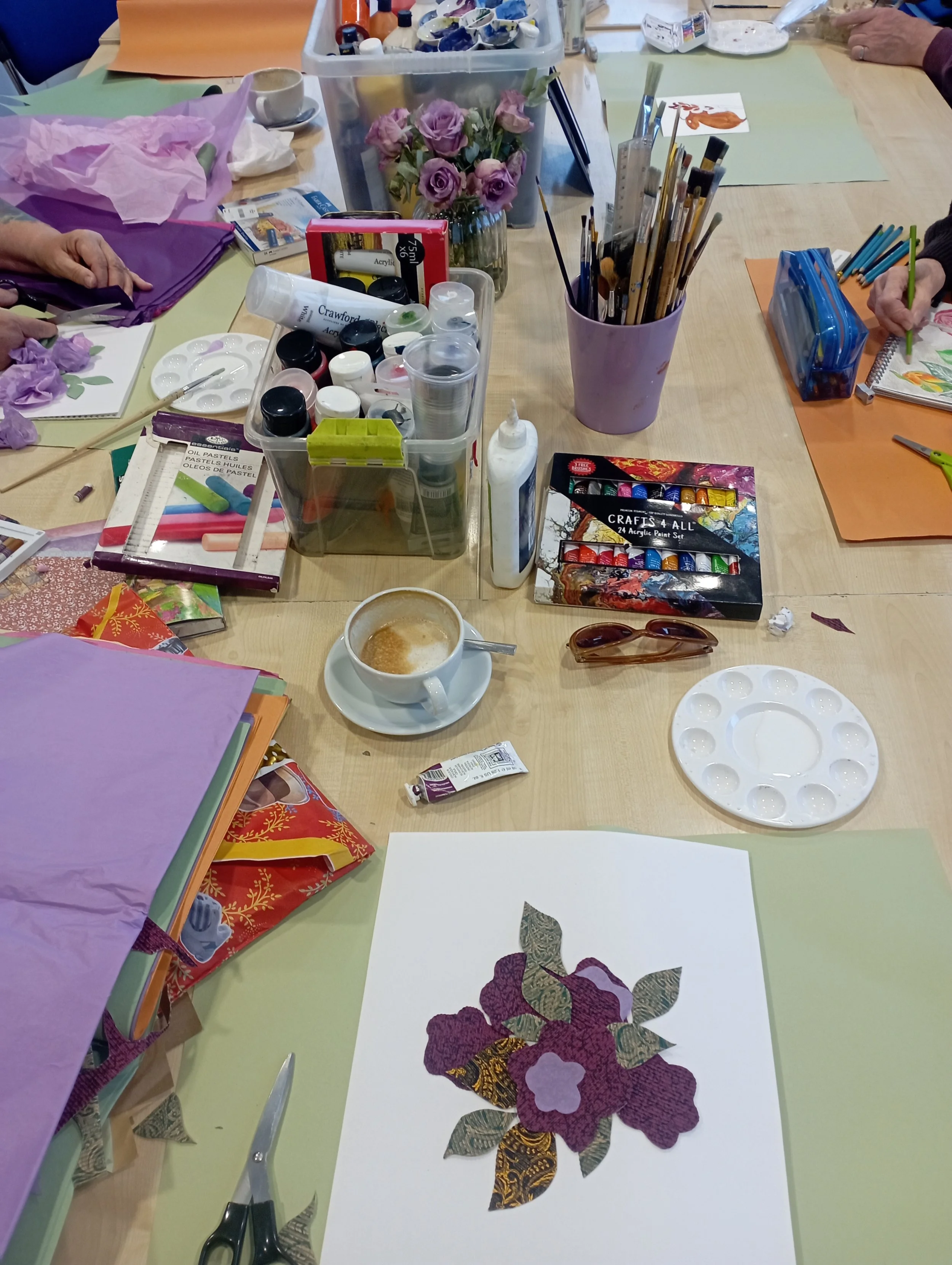 A crafting table with various art supplies including paints, brushes, scissors, and colored paper. There are also flowers, a cup of coffee, and a paper with a floral collage design.