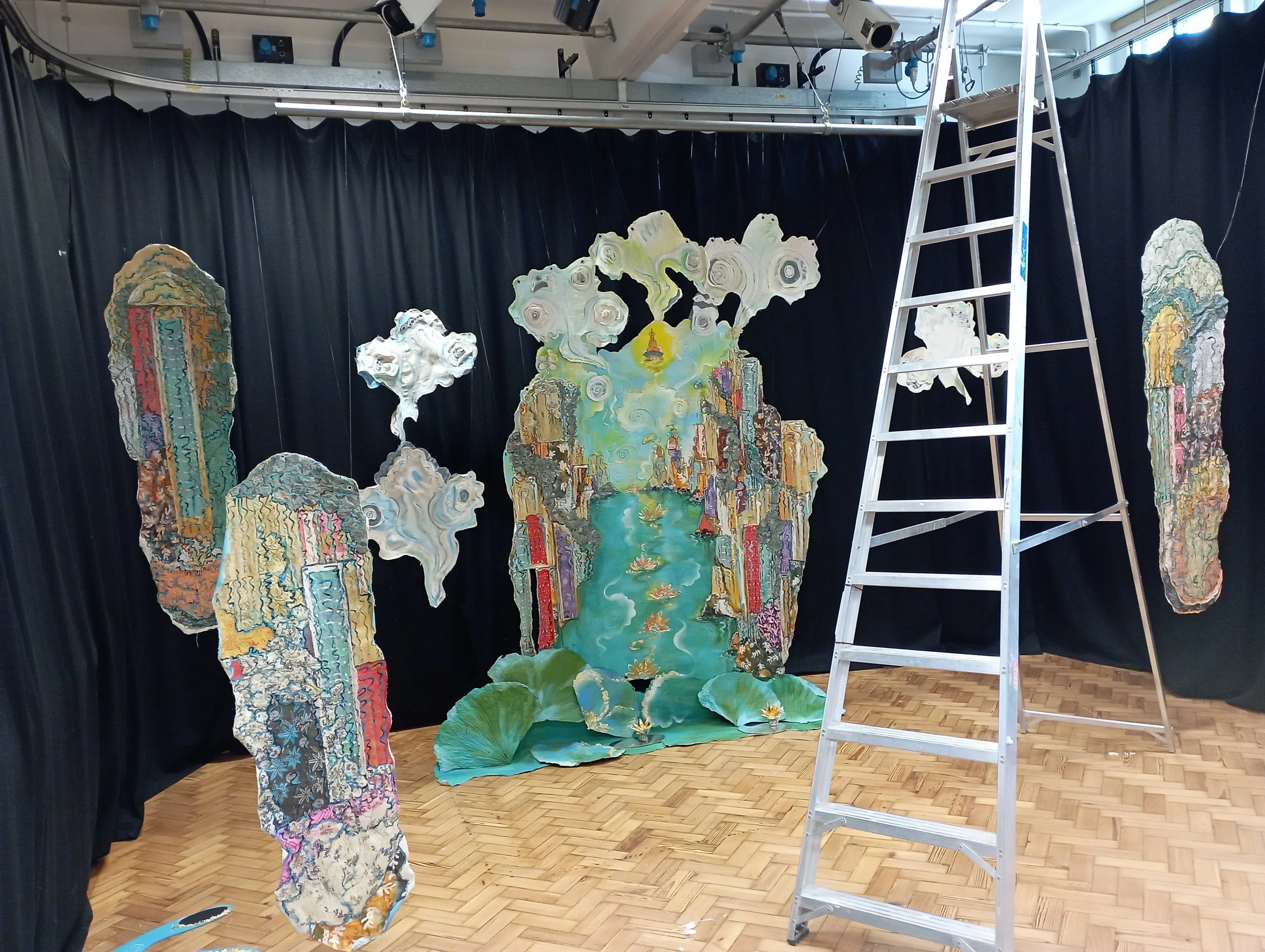 A colorful, artistic backdrop being assembled in a room with black curtains and wooden parquet flooring. The backdrop features abstract, layered shapes resembling a landscape with a river, buildings, clouds, and a yellow sun. A silver step ladder is 