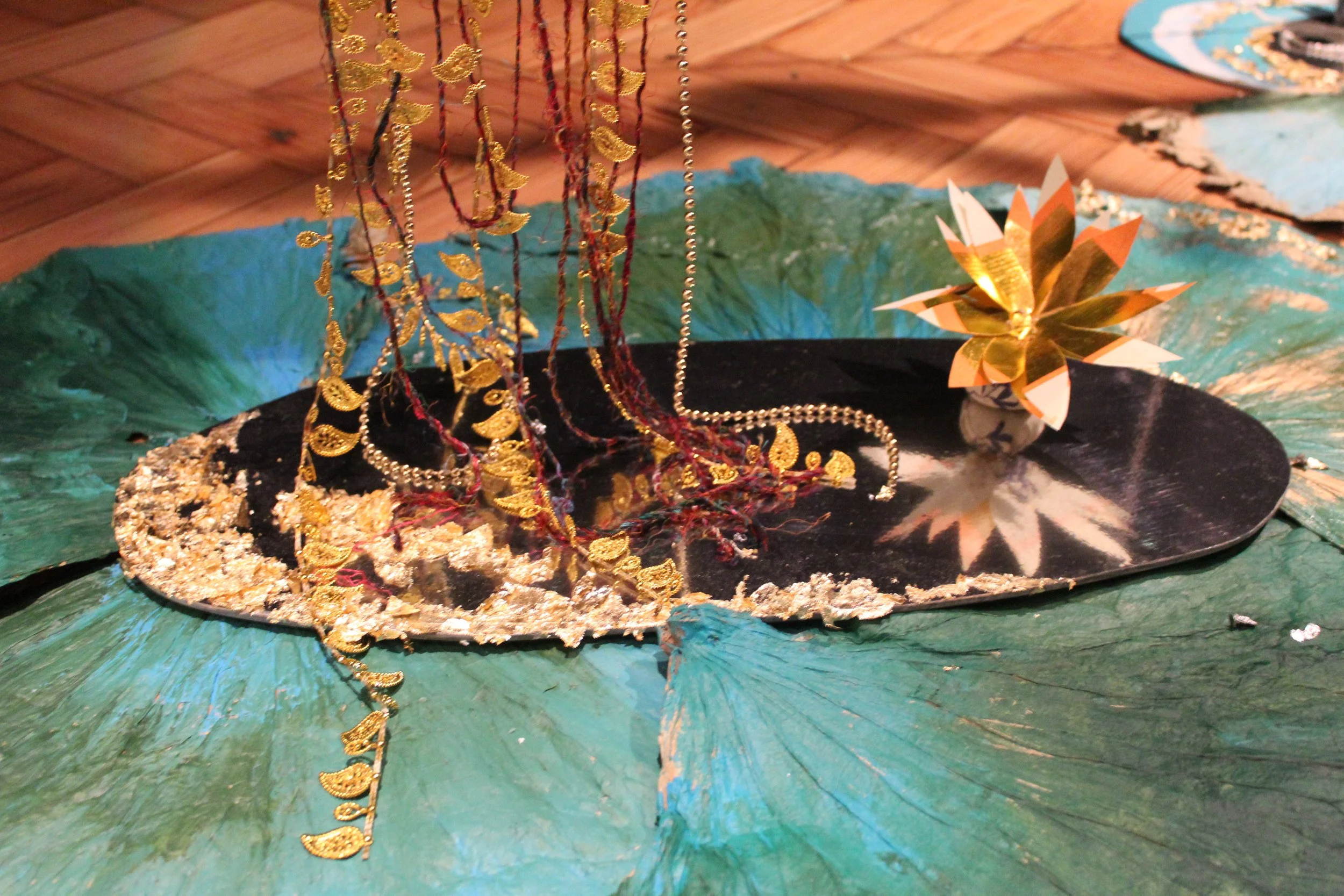 Mixed media art piece with green and black paper, gold and red decorative strands, a gold and white paper flower, and a reflective surface, possibly metallic or mirror, on a wooden floor.