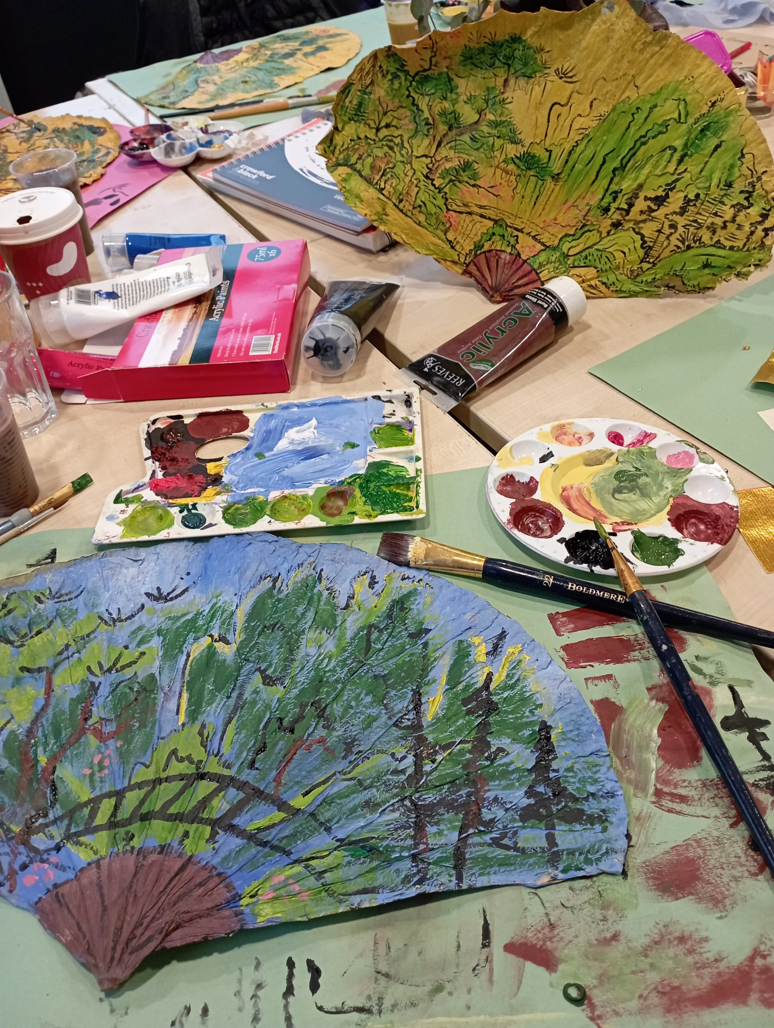 Colorful hand-painted folding fans depicting landscape scenes with trees and mountains on a cluttered art table with paint supplies.