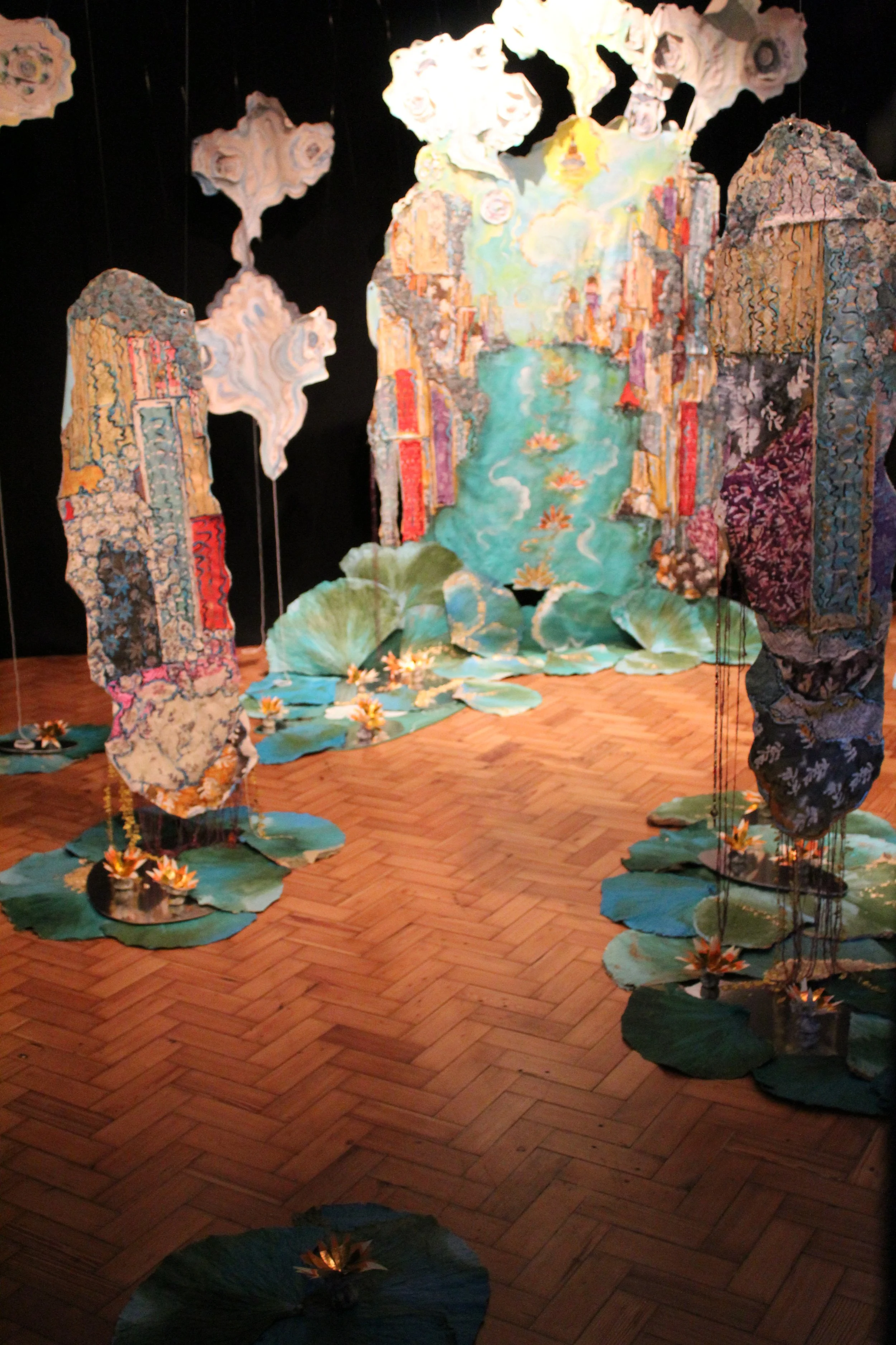 An art installation featuring large, decorative panels resembling layered fabric or tapestry with a nature scene, including a water body with lily pads and flowers, set on a wooden floor.