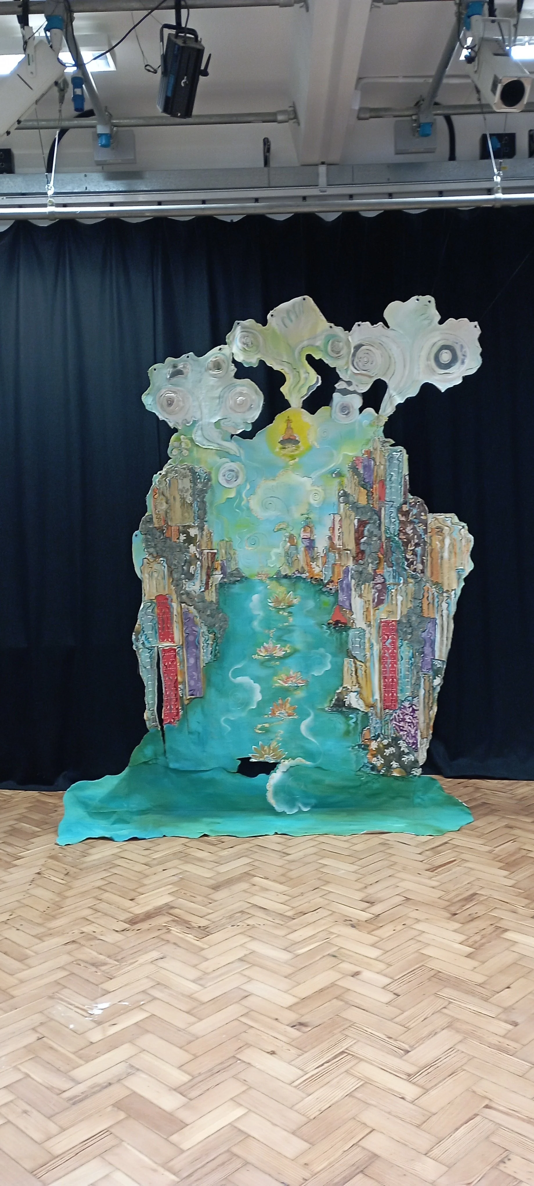 A painted theatrical backdrop with a landscape of tall, colorful buildings lining a river, topped by stylized clouds and a bright yellow sun in the sky, set against black curtains in an indoor space.