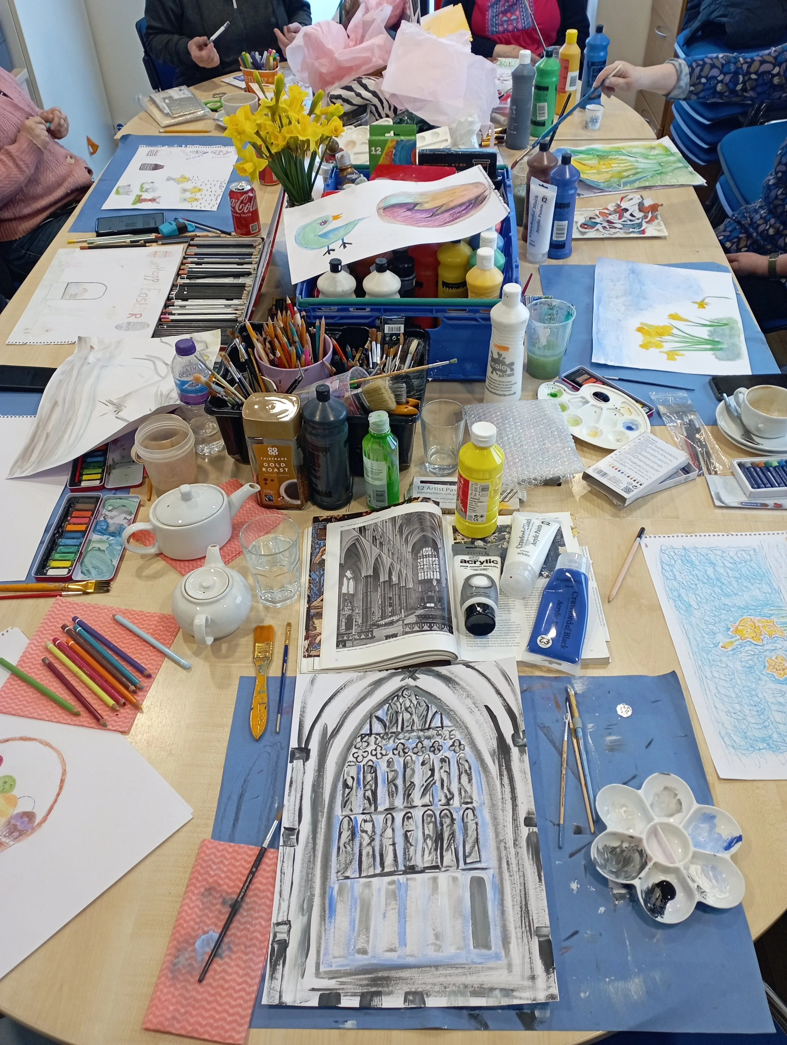 A table filled with art supplies, including watercolor paints, brushes, paper, and completed watercolor paintings, with people working on their art projects in the background.