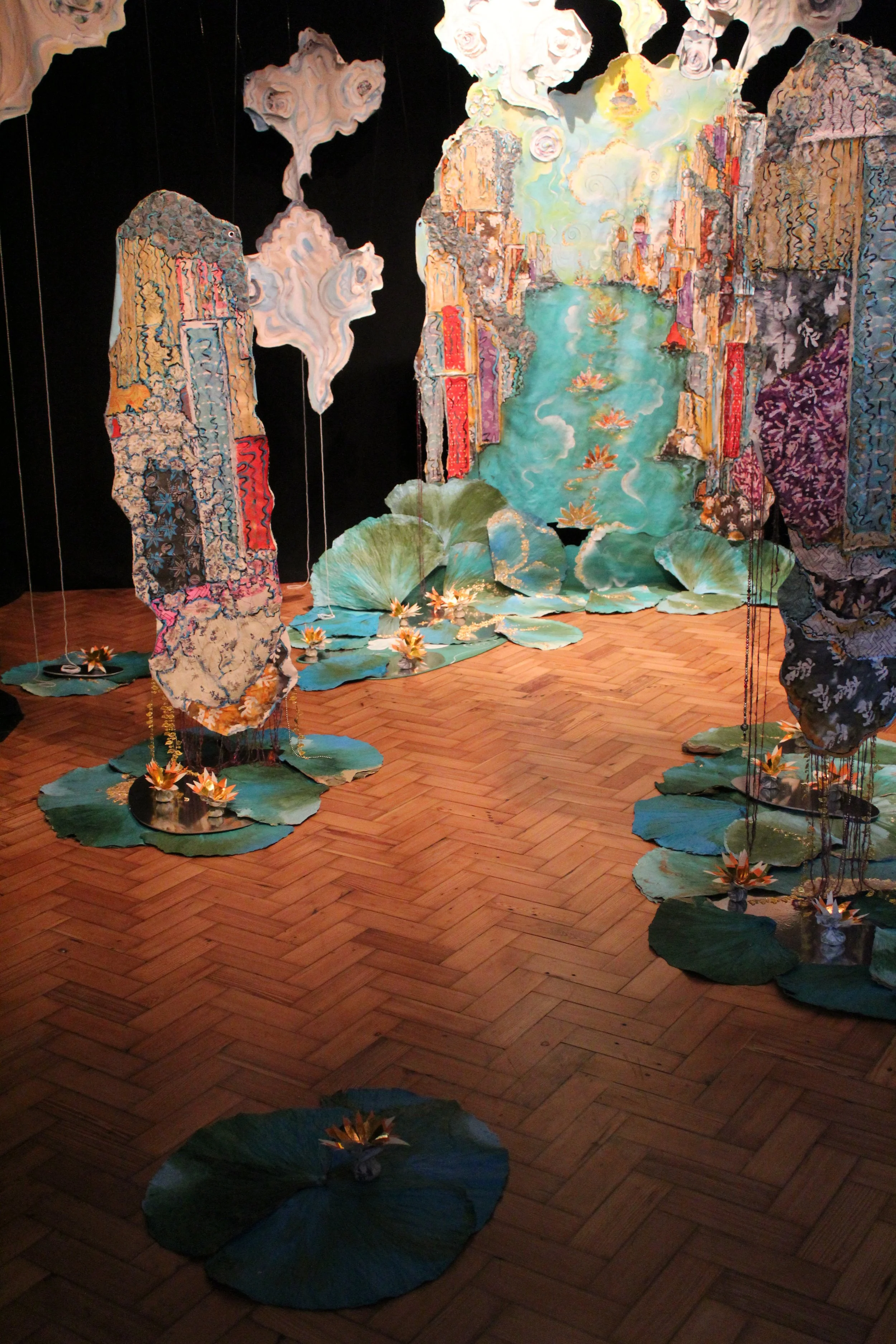 Art installation featuring painted paper cutouts of waterfalls, lily pads with lilies, and abstract clouds, creating a serene pond scene with a path of wooden flooring.